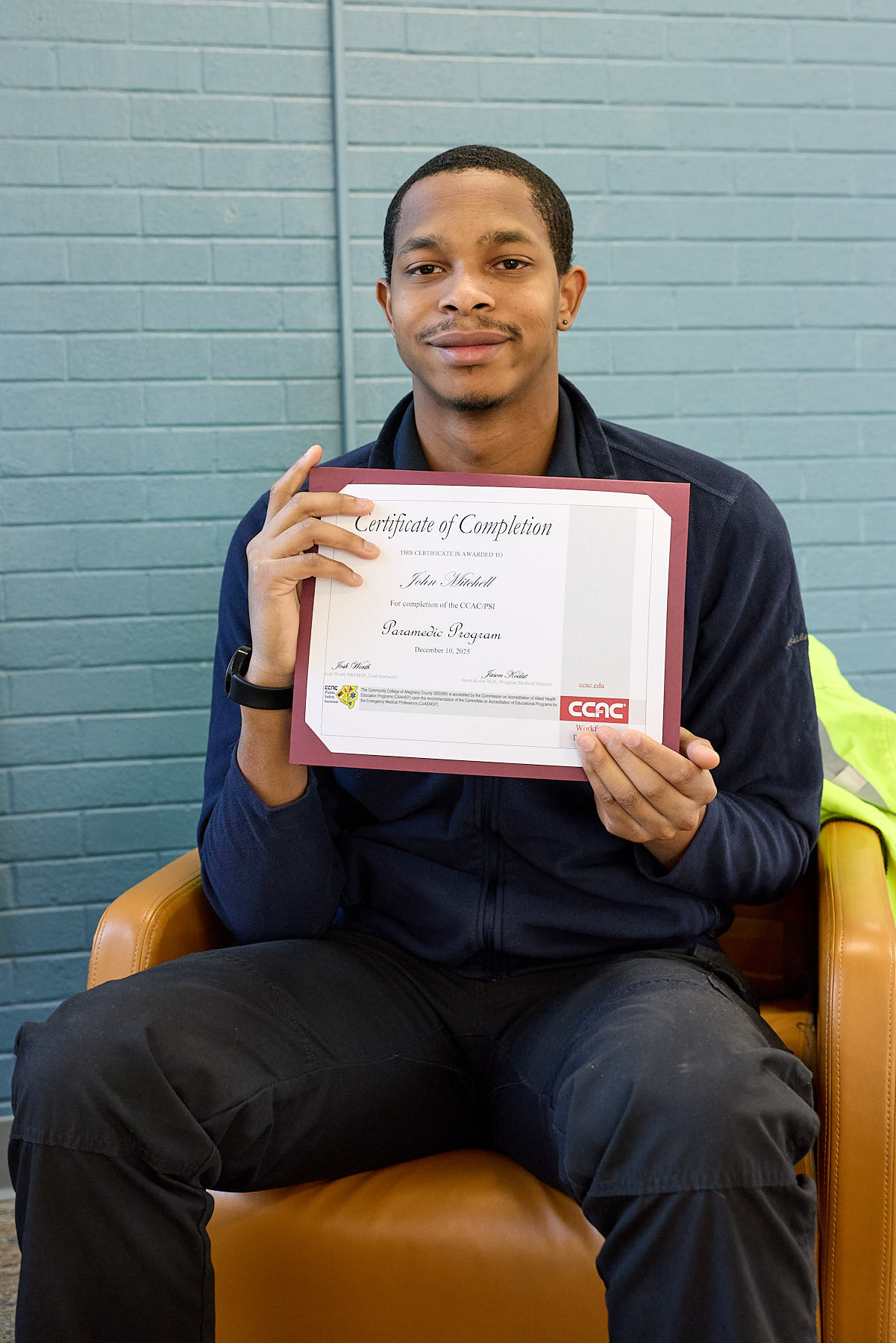 A man sitting in a chair holds up a certificate of completion from the Community College of Allegheny County, smiling at the camera.