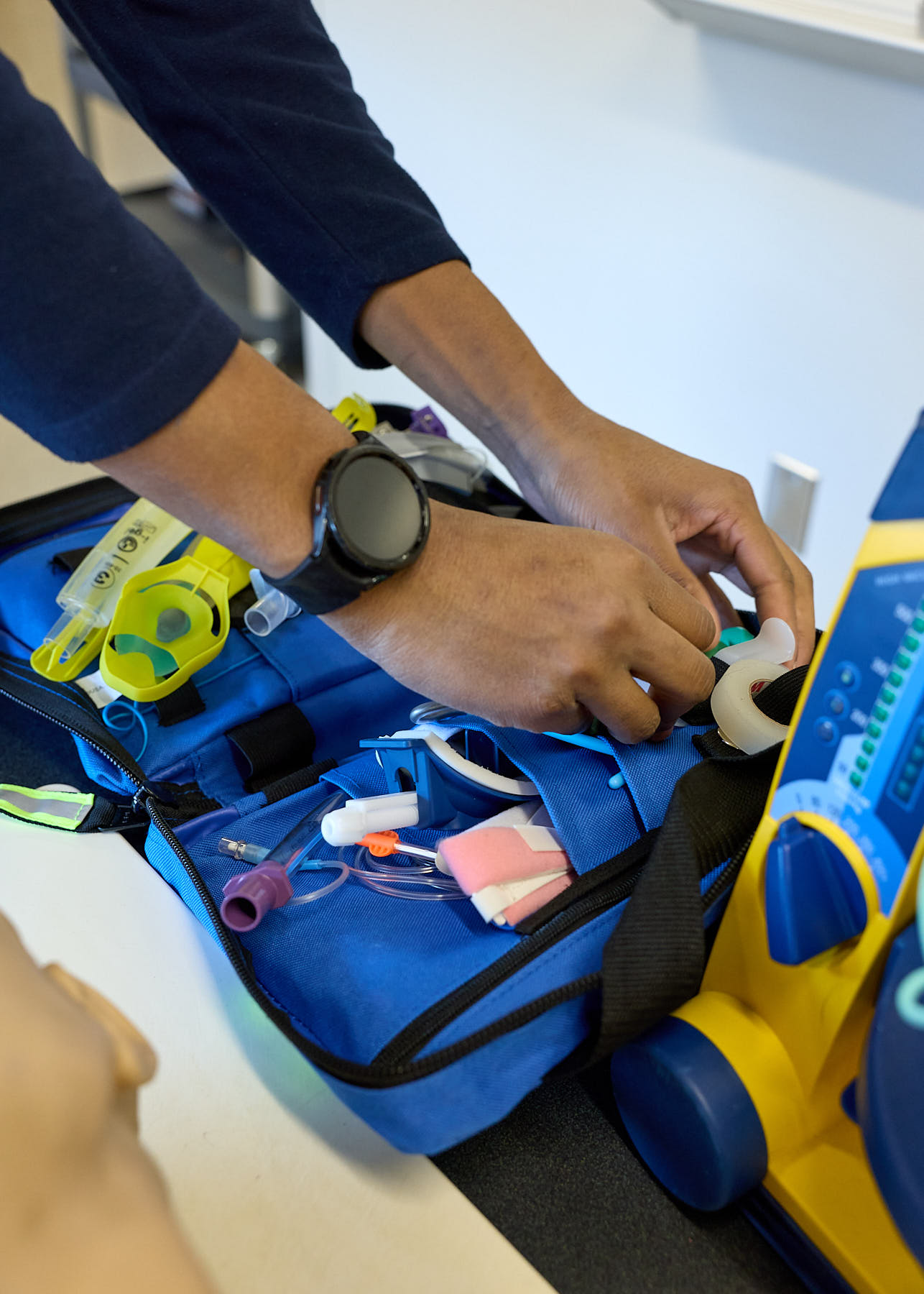 Person's hands organizing medical supplies in a blue emergency kit bag, with various tools and equipment visible, next to a medical device.