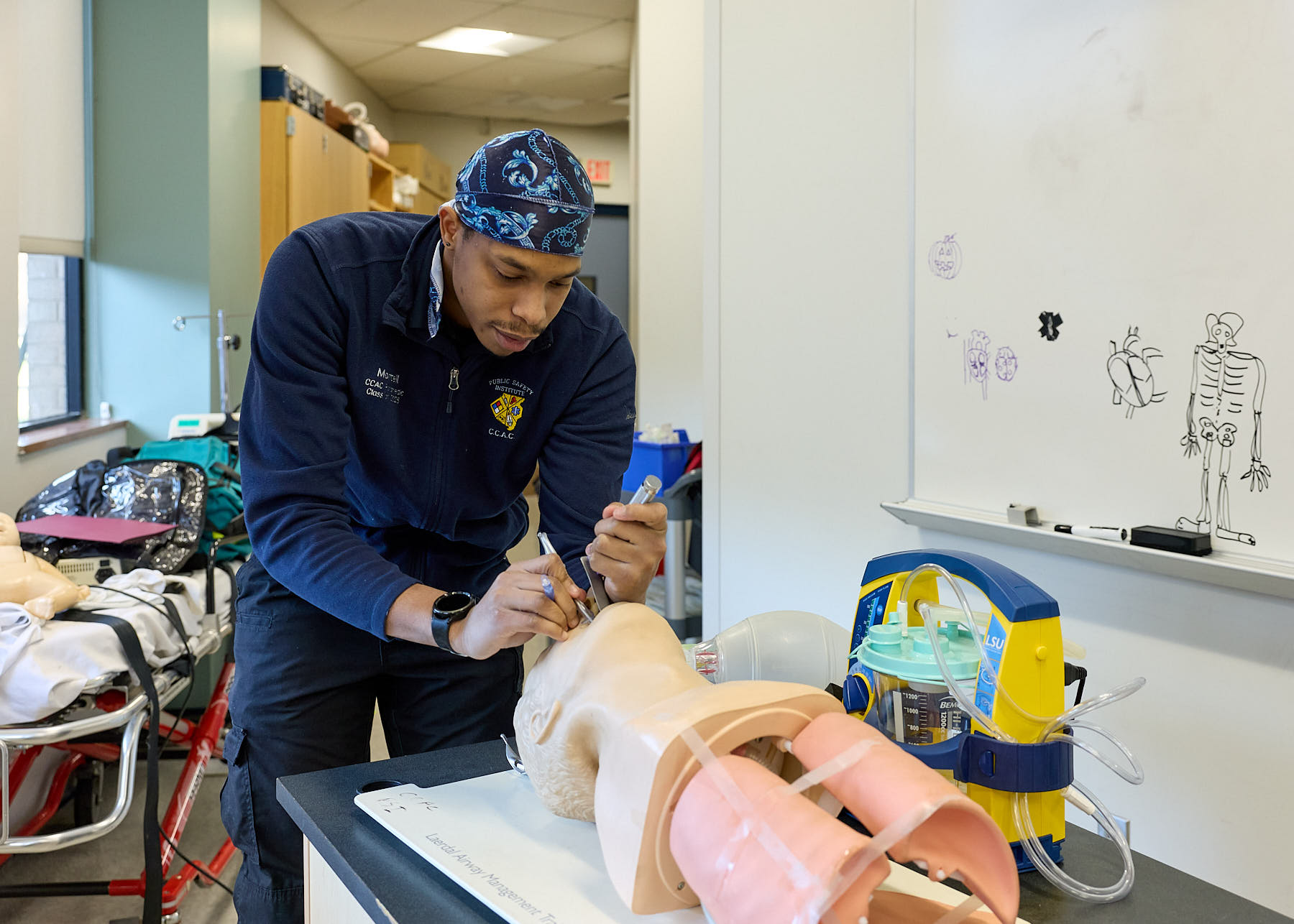 A man in medical attire practices a procedure on a medical mannequin in a classroom with medical equipment and anatomical drawings on a whiteboard.