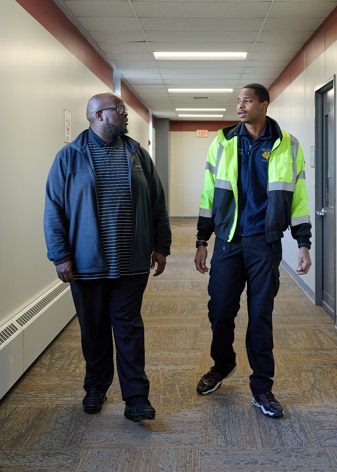 Two men walk side by side down a hallway, engaged in conversation. One wears a striped shirt and jacket, the other wears a hi-vis jacket over a dark uniform.