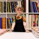 A person with glasses stands behind a table in a fabric store, surrounded by colorful bolts of fabric neatly arranged on shelves.
