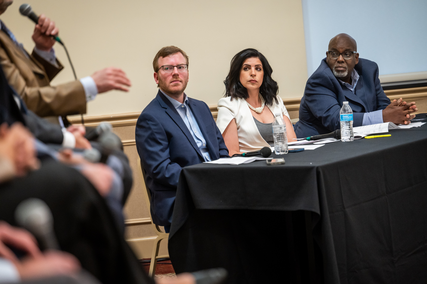Three panelists sit at a table listening to a speaker holding a microphone during a discussion event, with papers and water bottles on the table.