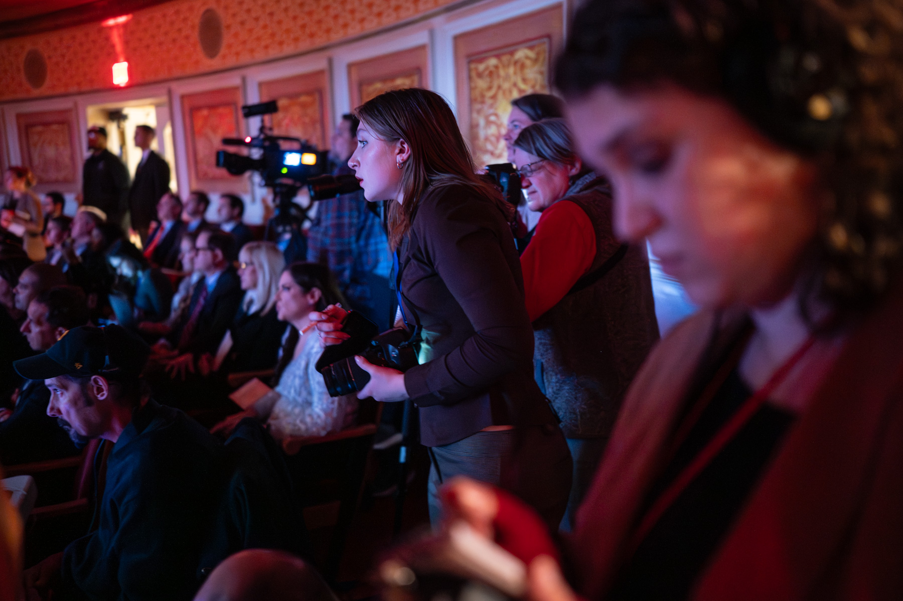 A group of people, including photographers and videographers, are standing and sitting in a dimly lit auditorium, focusing on an event happening out of frame.