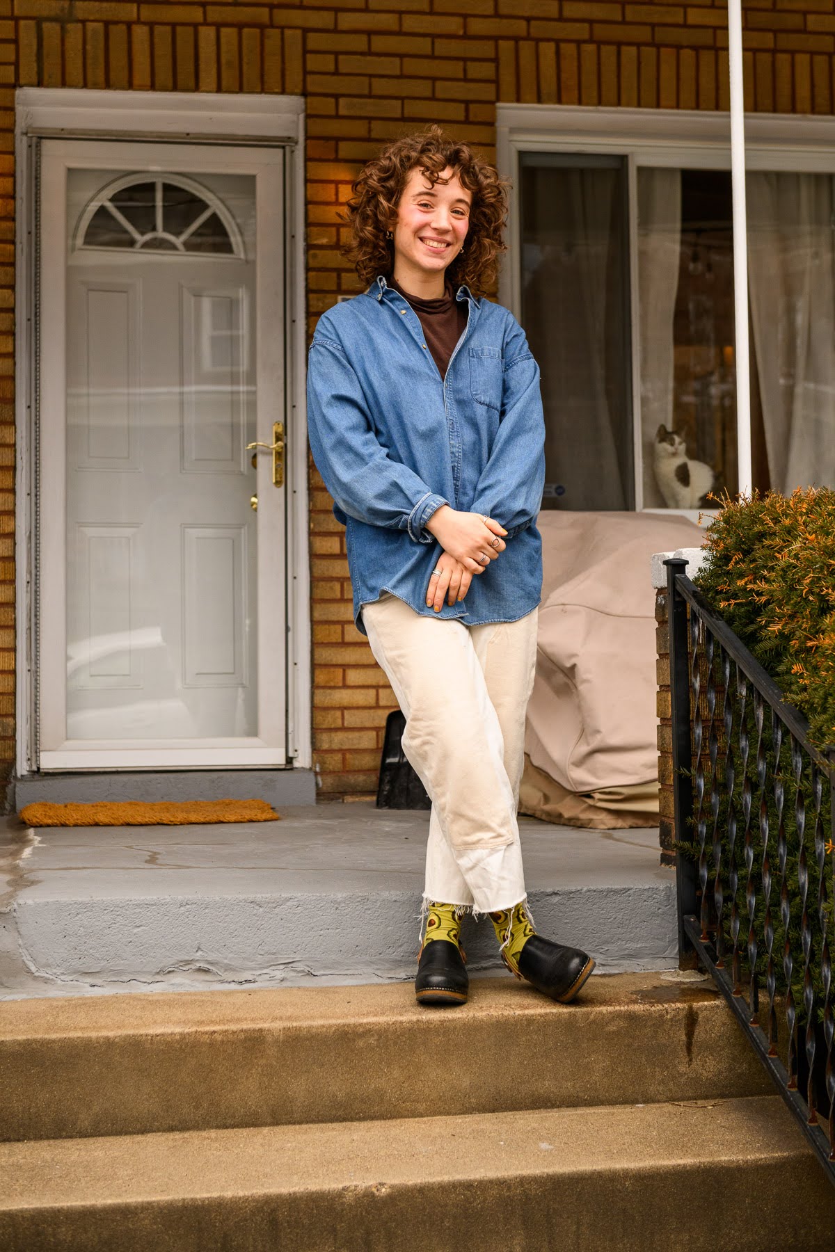 A person with curly hair stands on a porch, wearing a blue button-up shirt, light pants, patterned socks, and black shoes, smiling at the camera.