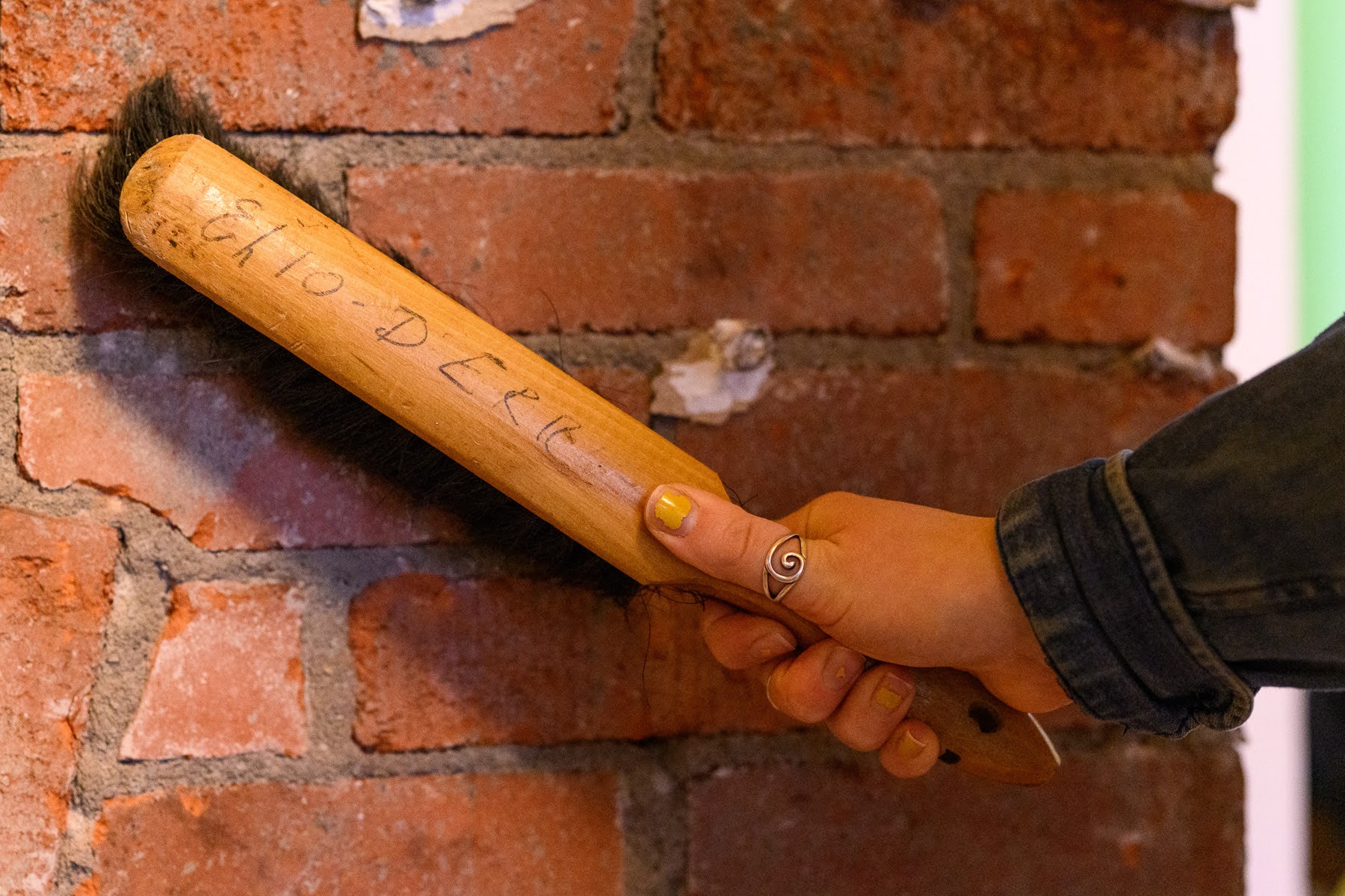A hand holding a wooden-handled brush with writing on it, brushing dust off a brick wall.