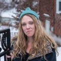 A woman in a teal hat and dark coat stands by an open wrought iron gate in a snowy residential neighborhood.