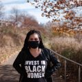A person stands outdoors on a path, wearing a black mask and a T-shirt that says "Invest in Black Women," with autumn trees in the background.