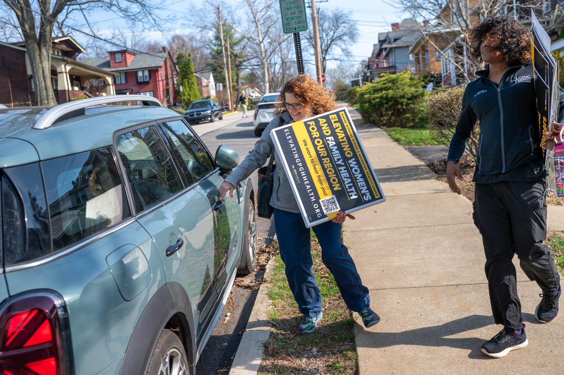 Two people on a sidewalk carry campaign signs supporting Magee-Womens nurses seeking public input in bargaining with UPMC; one is opening the door of a parked light blue car.
