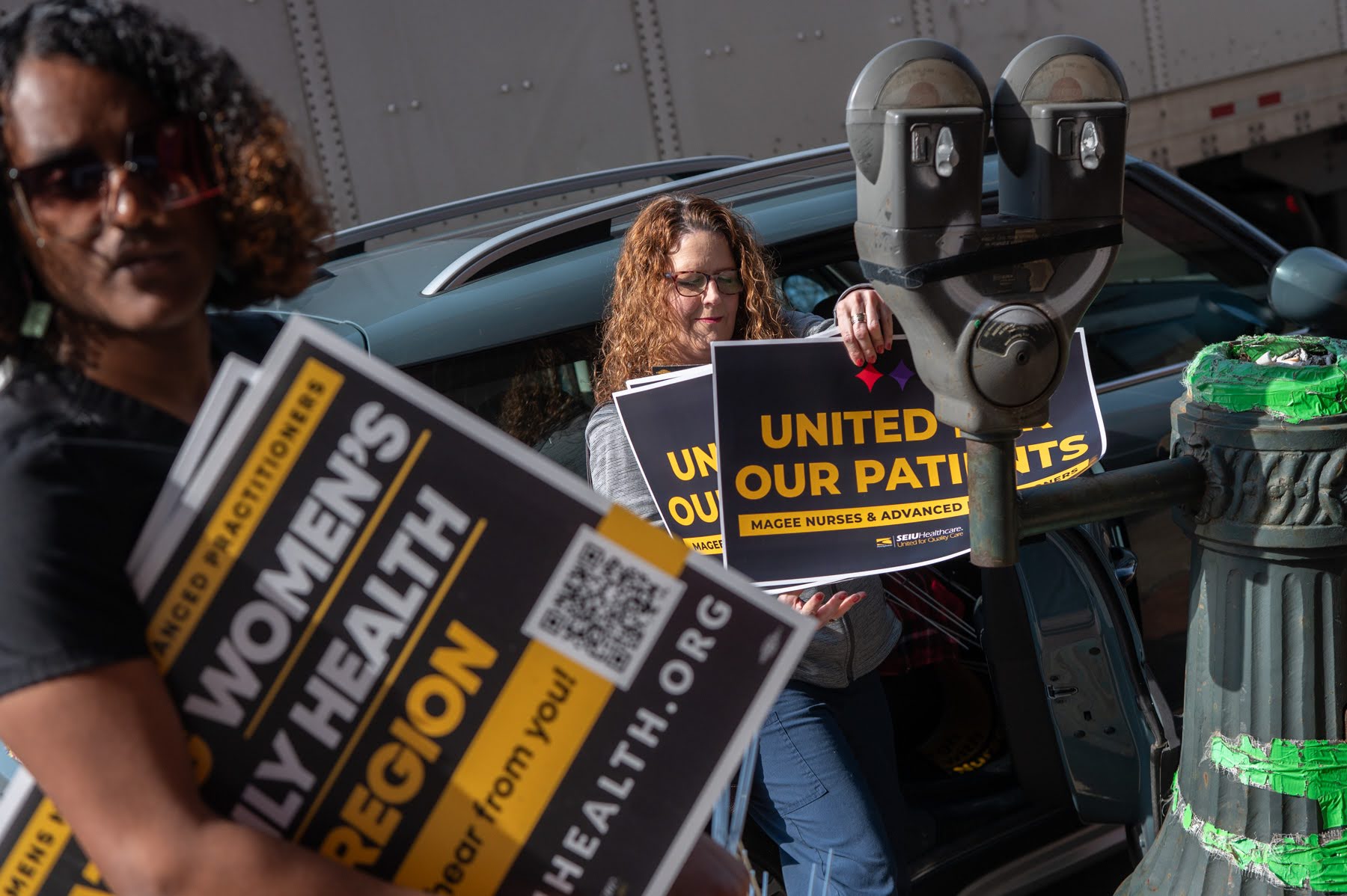Two people holding signs about women's health and patient care stand near a parked car on a city street.