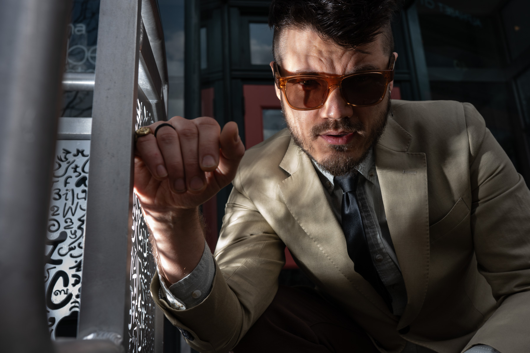 धूप का चश्मा और बेज रंग का ब्लेज़र पहने एक व्यक्ति कट-आउट पैटर्न वाली एक धातु संरचना के पास आगे की ओर झुका हुआ है, जिसके चेहरे पर सूरज की रोशनी की छाया पड़ रही है।