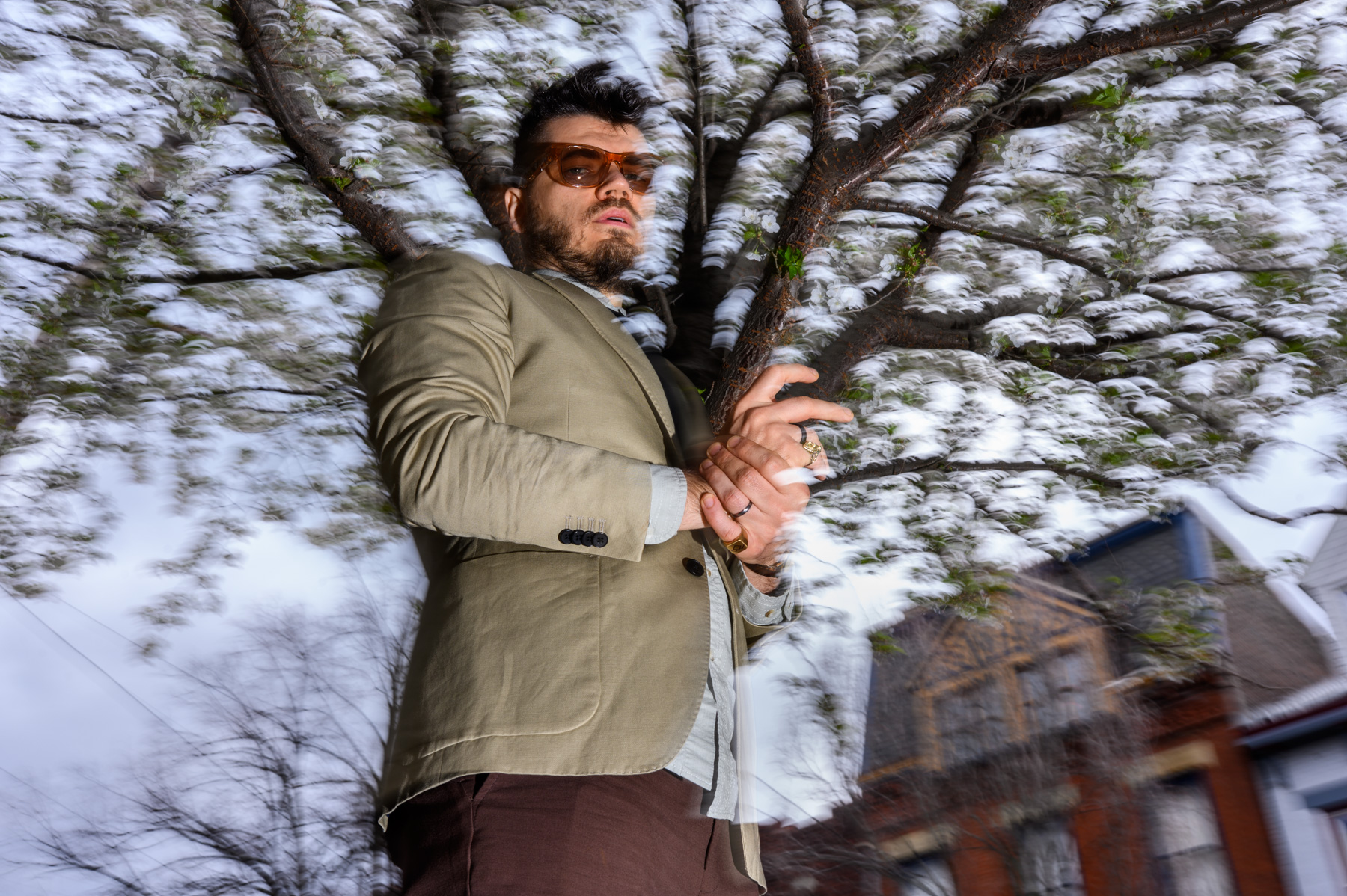 धूप का चश्मा और ब्लेज़र पहने एक आदमी एक खिले हुए पेड़ के सामने खड़ा है, जिसकी पृष्ठभूमि में इमारतें दिखाई दे रही हैं।