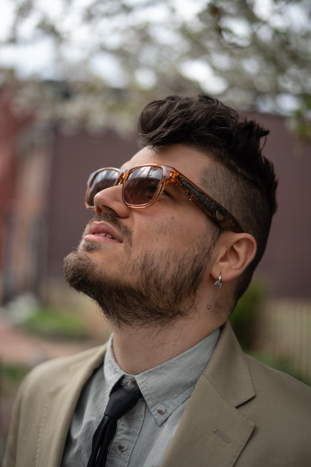 छोटे, सजे हुए बाल और दाढ़ी वाला एक व्यक्ति, धूप का चश्मा, टैन सूट, हल्की शर्ट और काली टाई पहने हुए, बाहर की ओर देख रहा है।