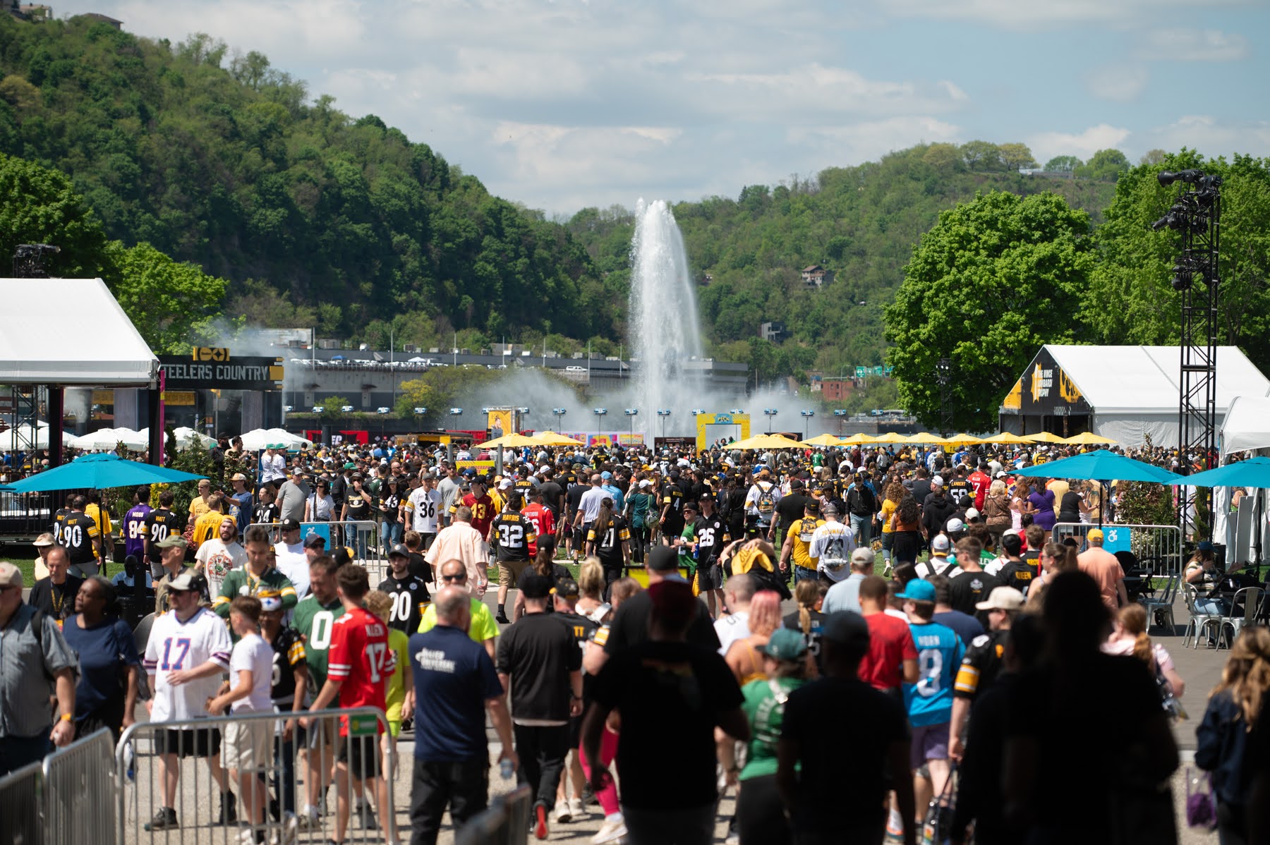 A large crowd of people, many in sports jerseys, gather outdoors on a sunny day with a fountain and tree-covered hills in the background.