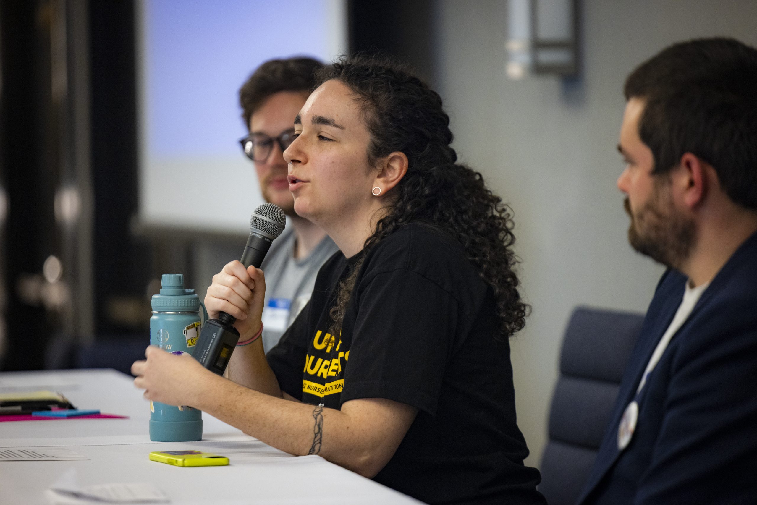 Person speaking into a microphone while seated at a table with two others at a panel discussion; water bottle and papers visible on the table.