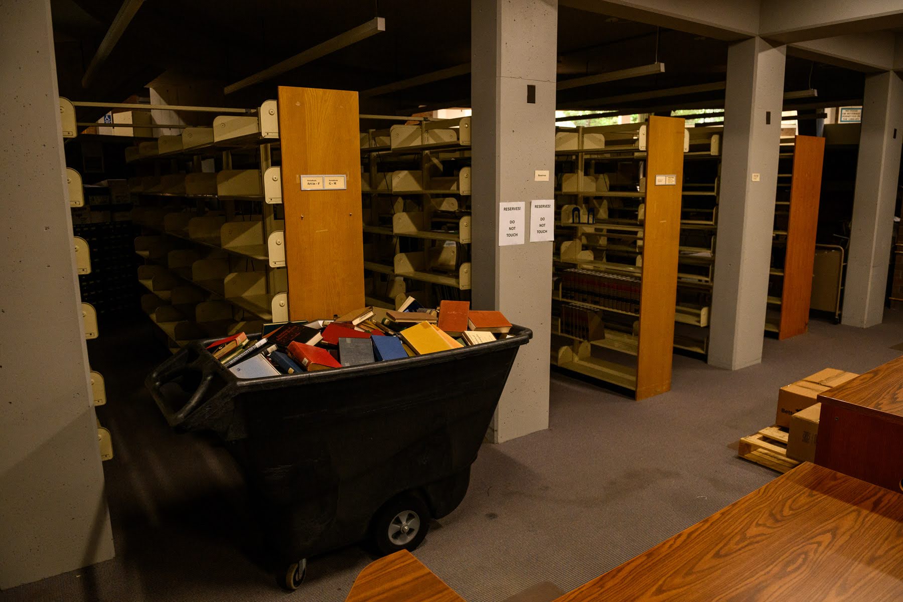 A large cart filled with assorted books sits in a dimly lit library storage area with mostly empty bookshelves and some wooden tables.