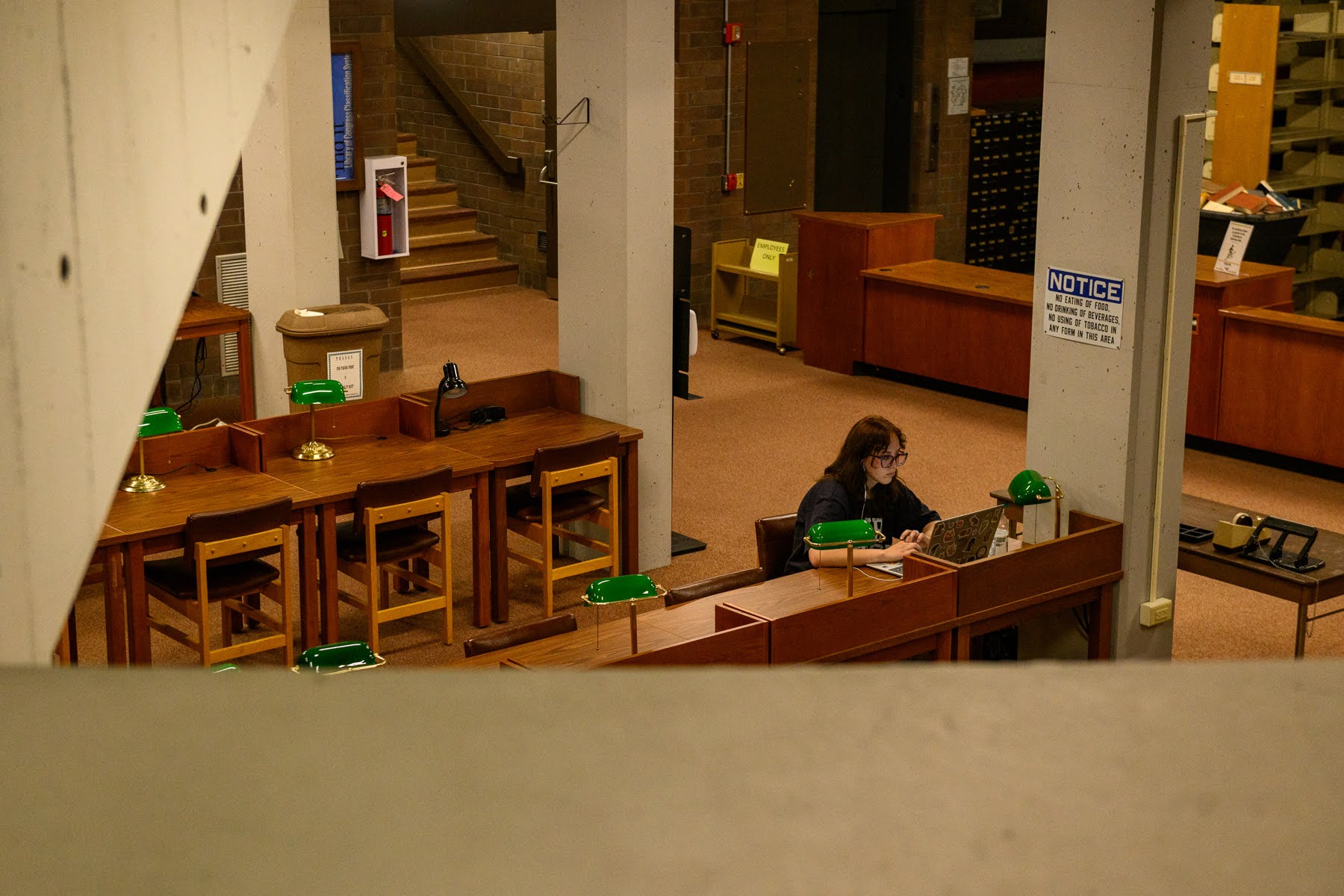 A person sits alone at a desk with a laptop in a library, surrounded by empty wooden tables and green-shaded lamps.