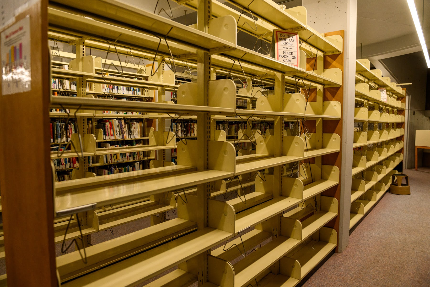 Rows of mostly empty yellow library shelves with some books on the shelves in the background; signs instruct users to place books on carts.