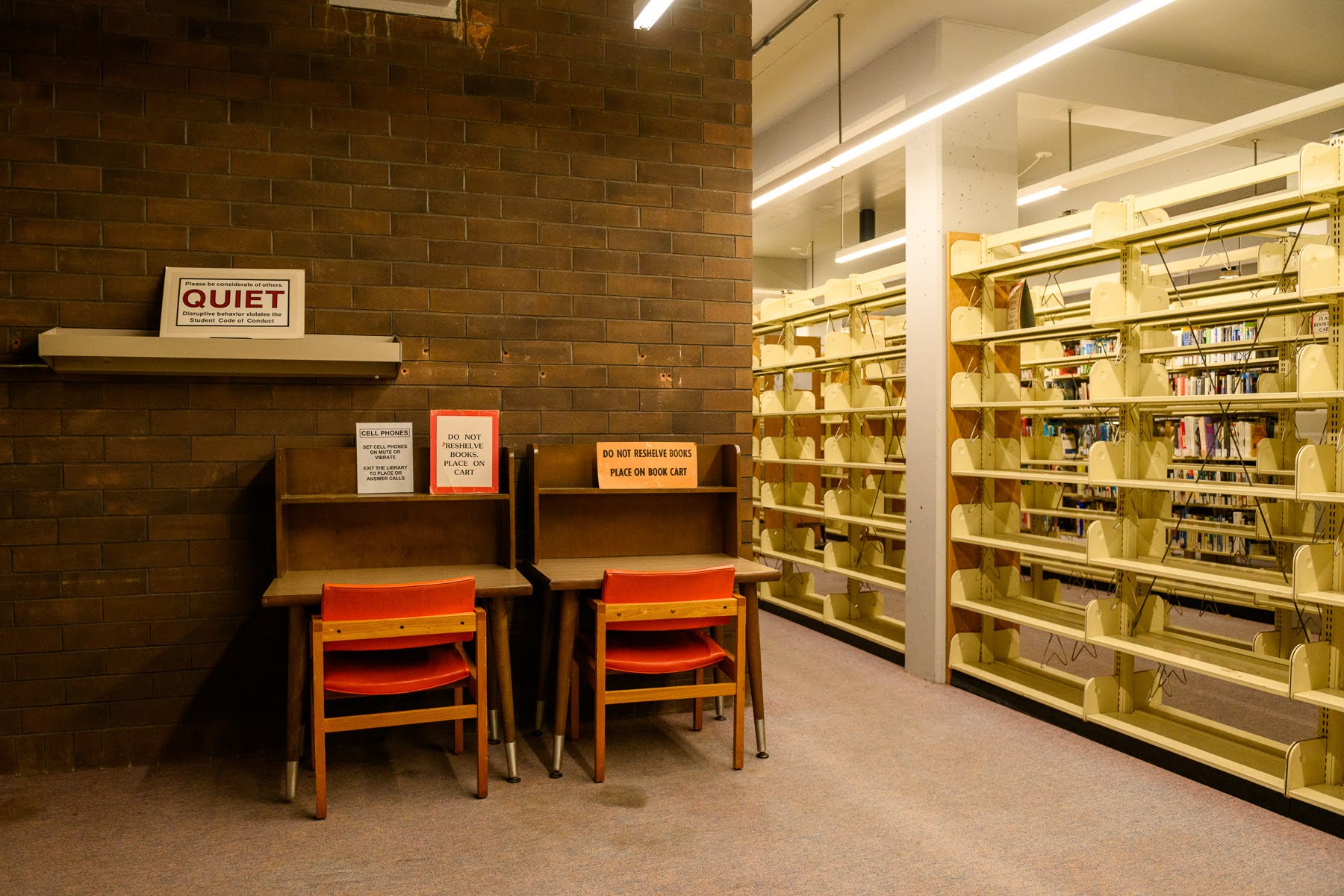 Two study desks with orange chairs are placed against a brown brick wall with a 