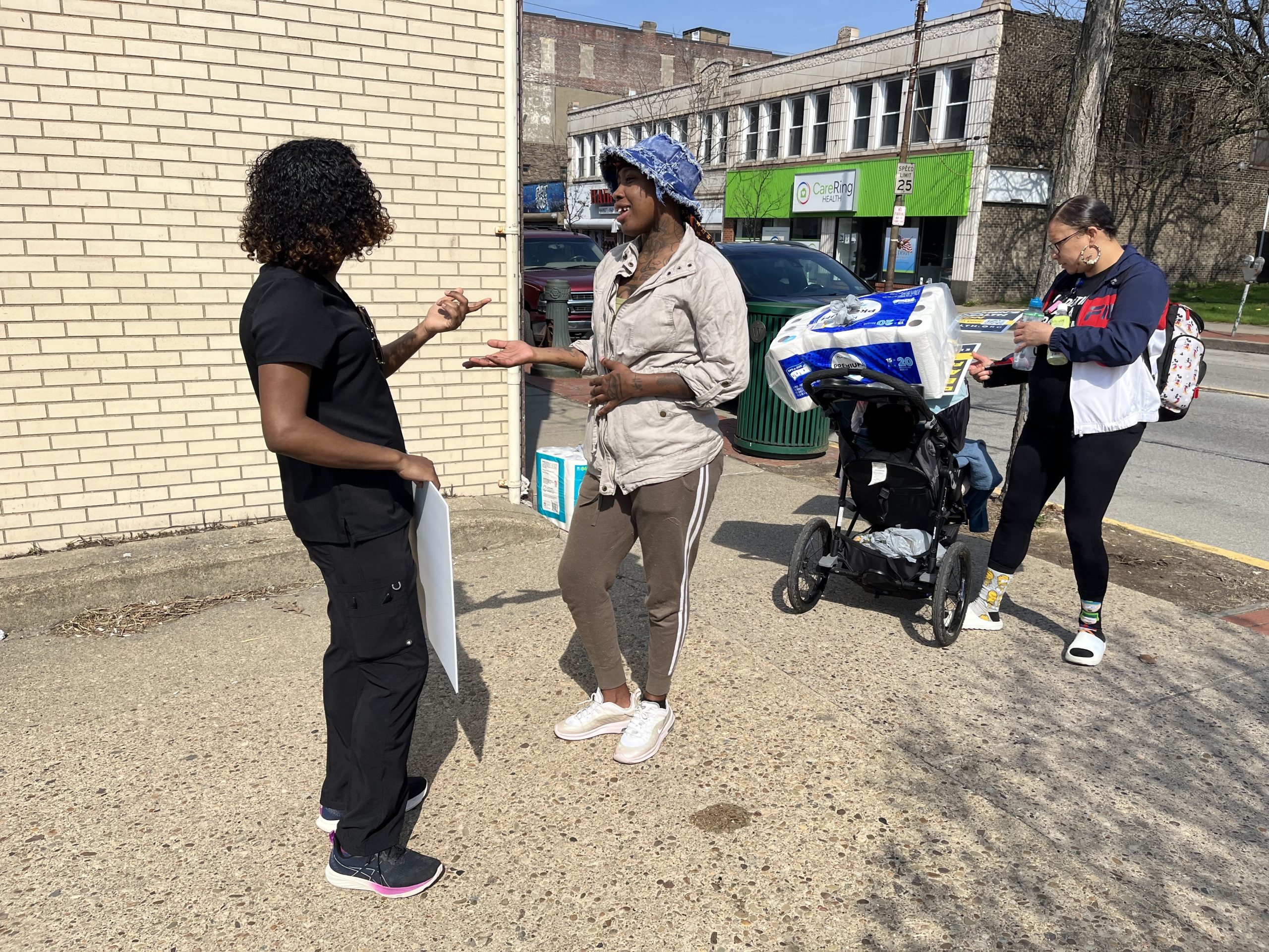 Two people have a conversation on a sidewalk near a brick building, while another person walks by pushing a stroller and carrying paper towels—just steps away, Magee-Womens nurses seek public input in bargaining with UPMC.