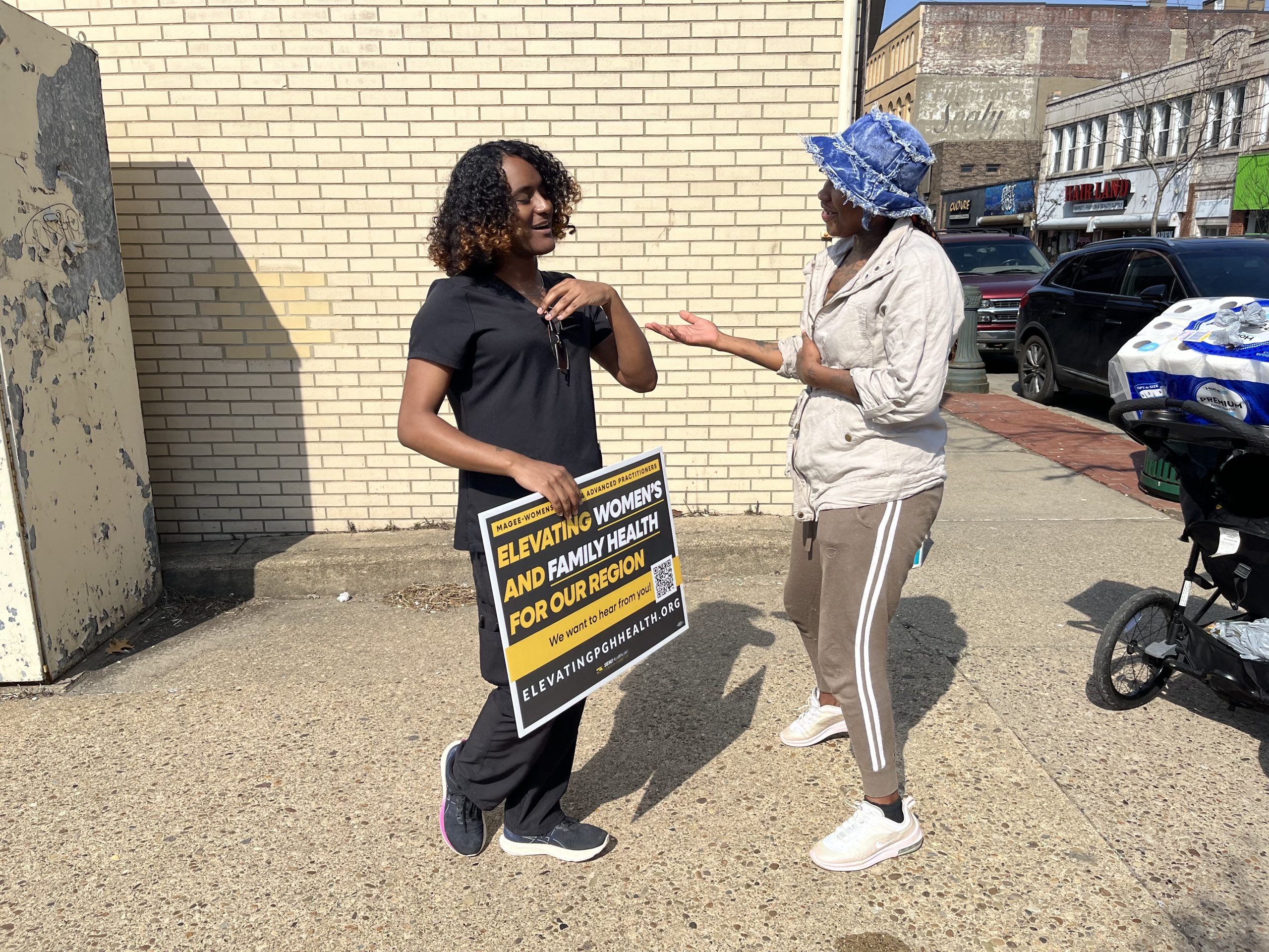 Two women stand talking on a sidewalk; one holds a sign that reads 