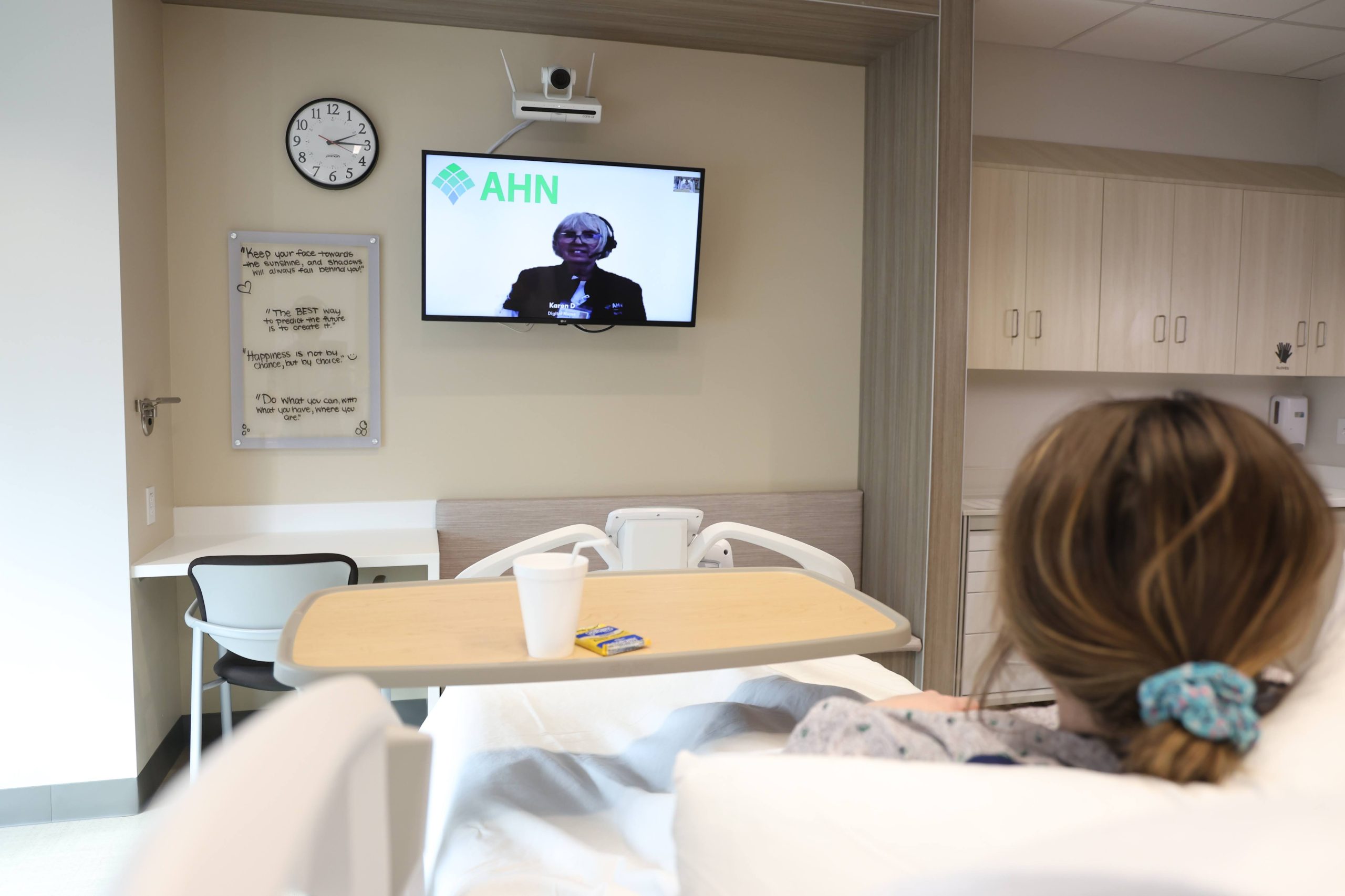 A patient lies in a hospital bed watching a telemedicine consultation with an AHN doctor on a wall-mounted TV screen.