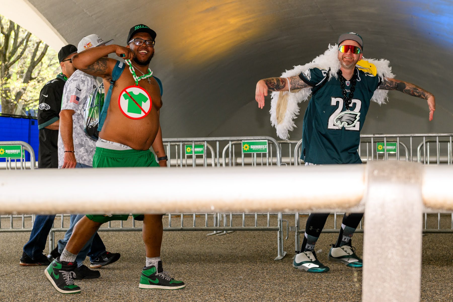 Two people dressed in Philadelphia Eagles gear pose playfully near a barrier; one wears large white wings and sunglasses, the other points while holding his shirt open.