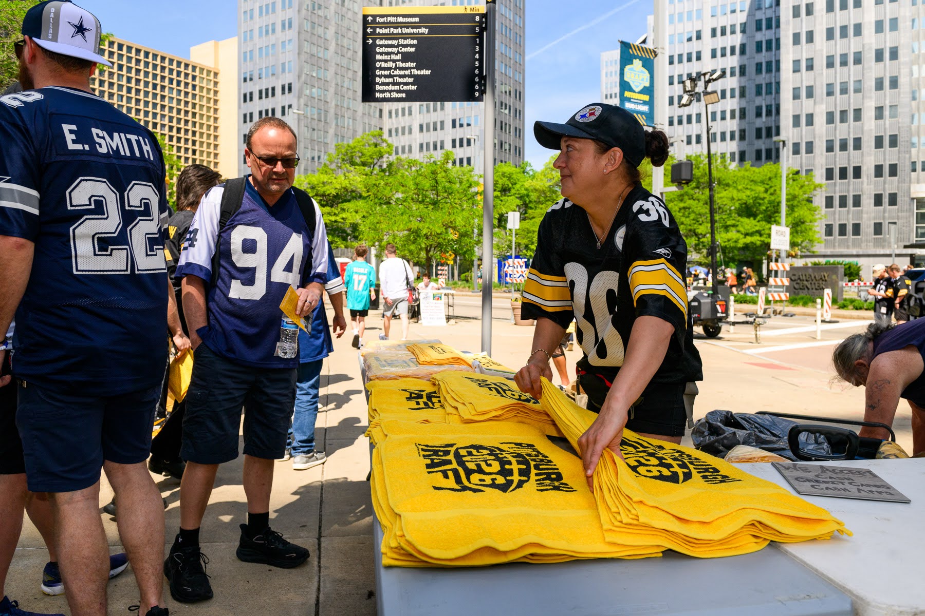 A person in a Steelers jersey arranges yellow towels on a table outside, while people in Cowboys jerseys stand nearby in a city setting.
