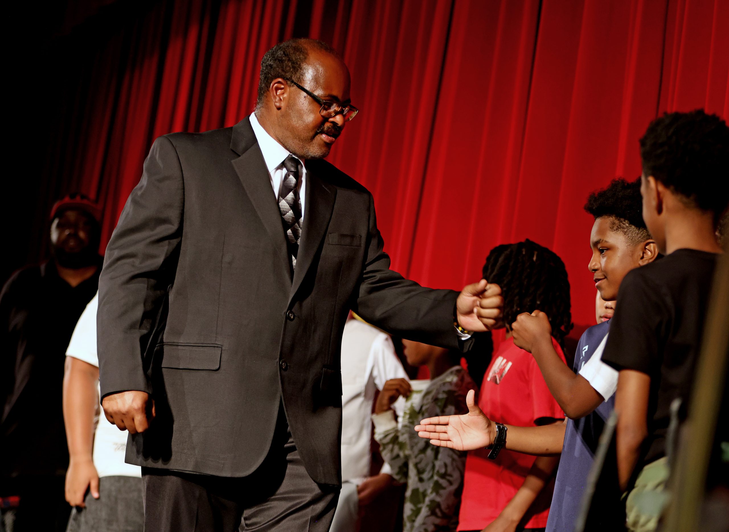 A man in a suit gives a fist bump to a boy on stage, with several other children standing nearby in front of a red curtain.