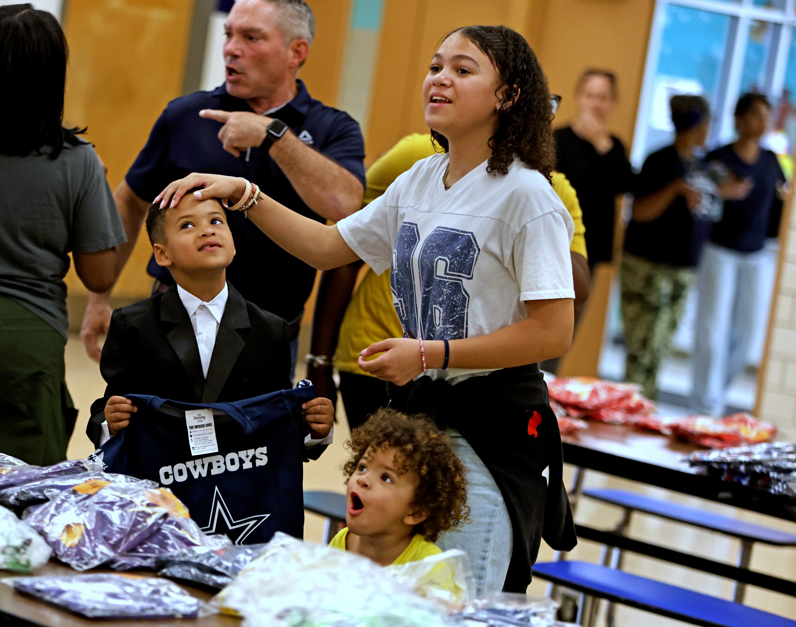 Three children stand by a table with clothing items, one holding a Cowboys shirt, while an adult gestures in the background inside a school cafeteria.