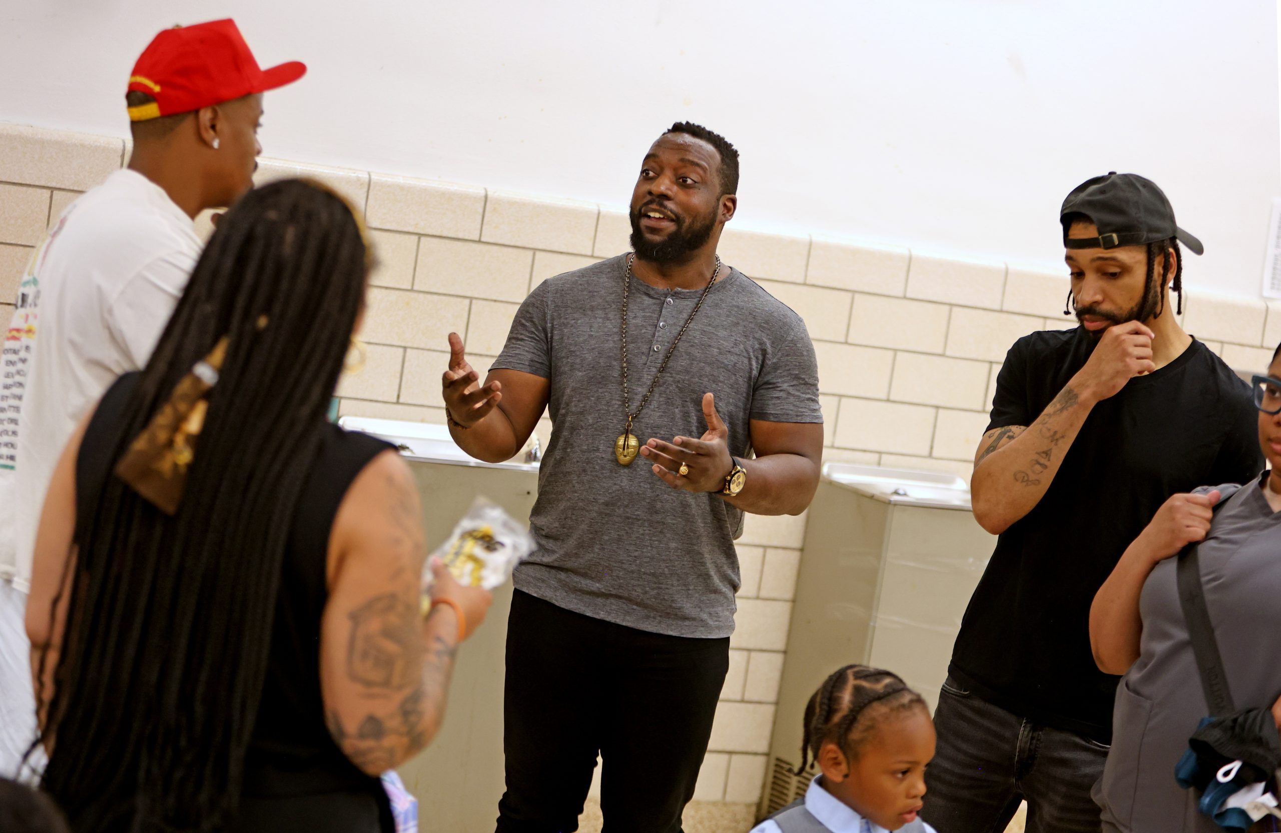 A group of adults stands and talks in a school hallway with children nearby. One man gestures while speaking; others listen.