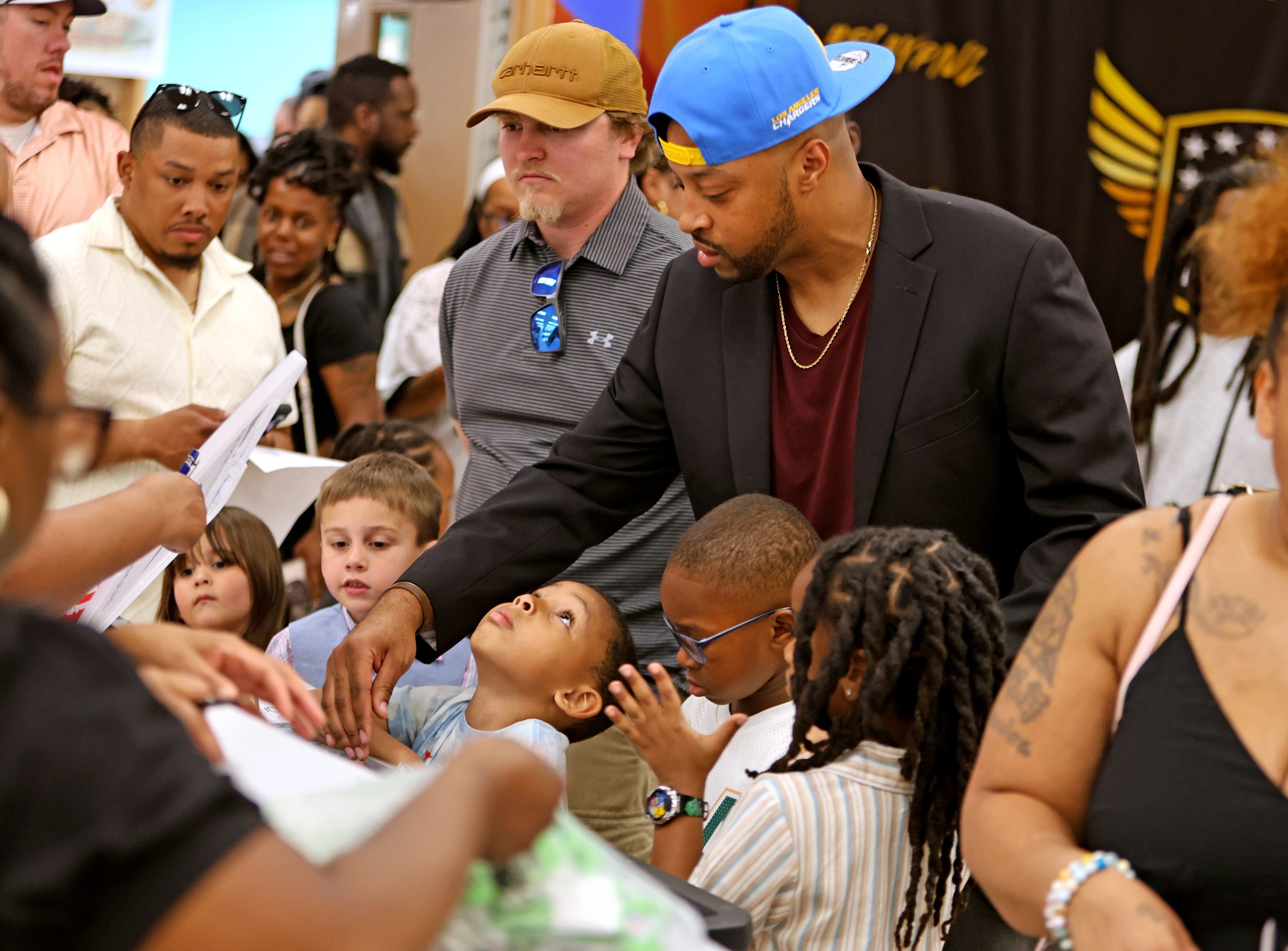 Adults and children gather around a table indoors, with one man in a blue cap and blazer reaching toward the children. The scene appears busy and interactive.
