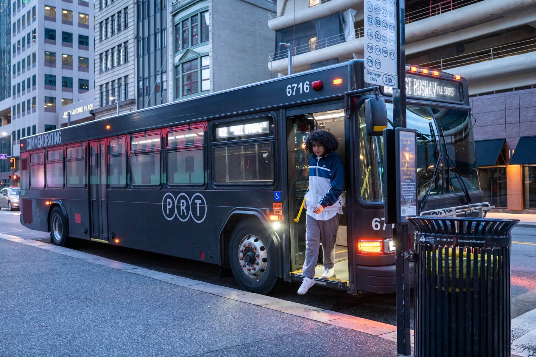 A person steps off a PRT city bus onto a sidewalk in an urban area at dusk, with buildings and streetlights in the background.