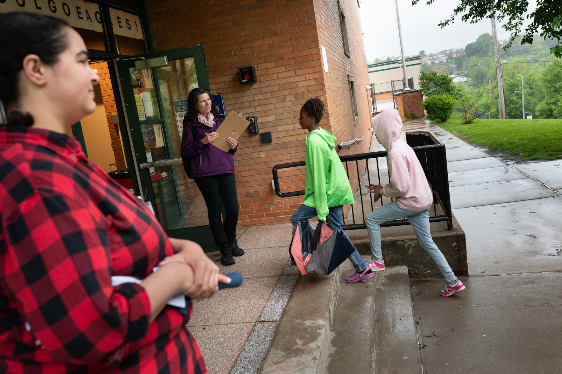 Two children walk up school steps while a woman holding a clipboard greets them at the door; another adult stands nearby on a rainy day.