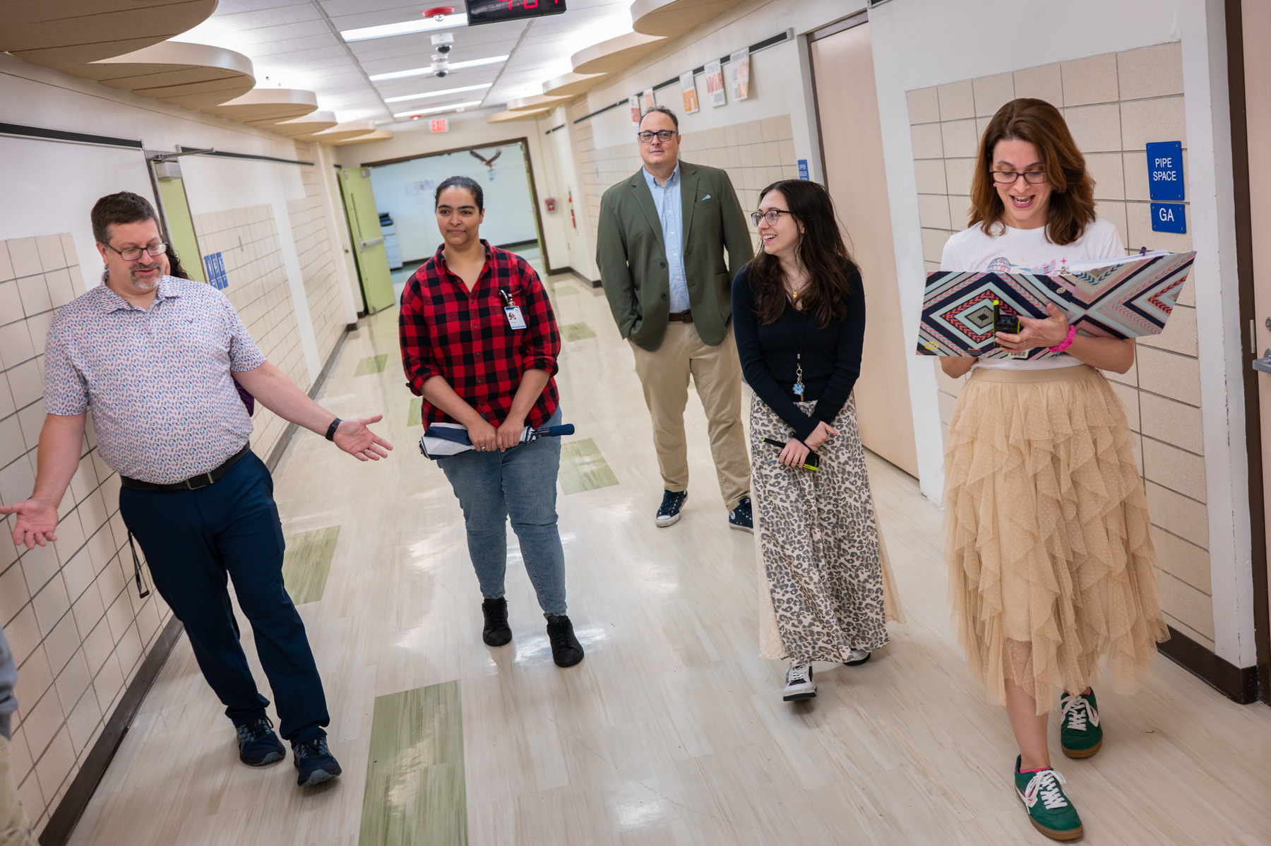 Five adults walk and talk in a school hallway, with one woman reading from a colorful folder while others listen.