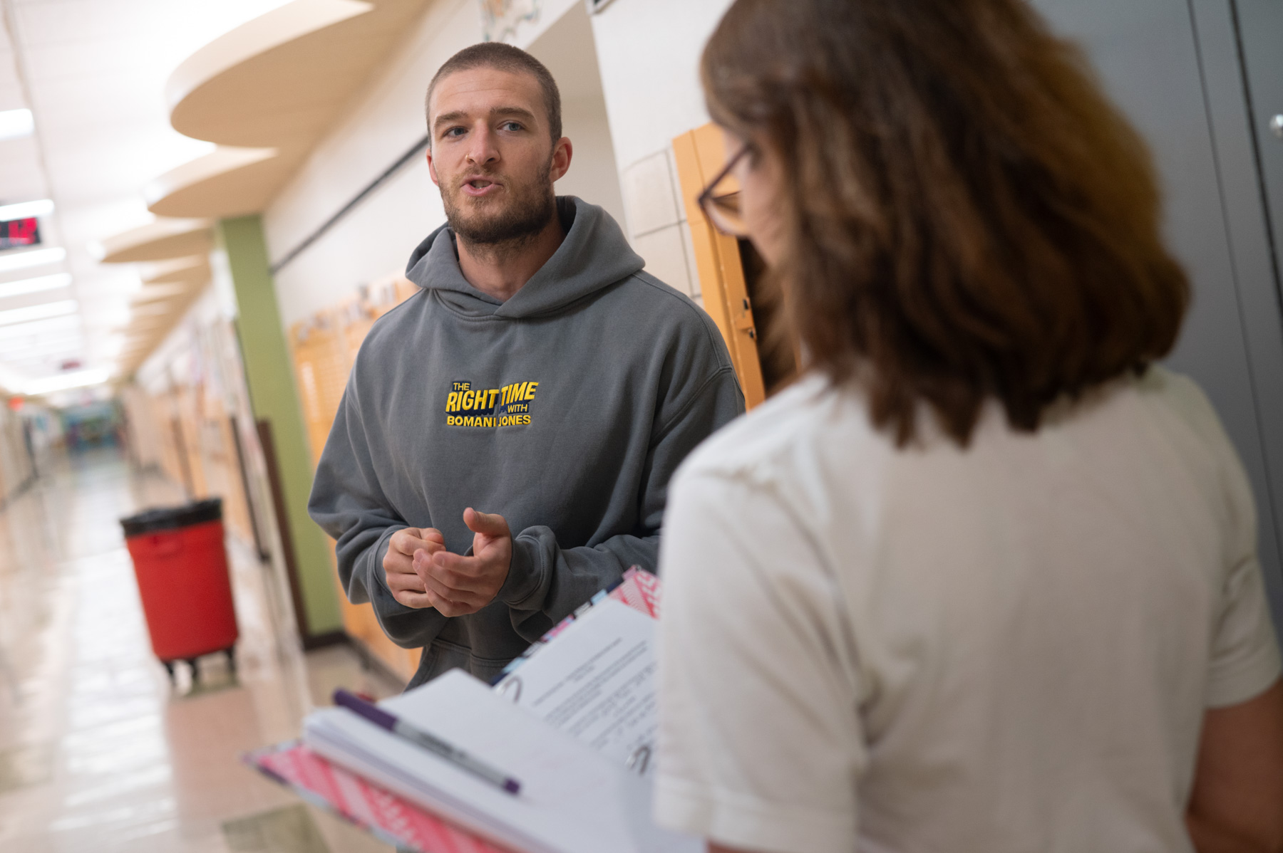 A man in a gray hoodie talks to a woman holding a folder in a school hallway lined with lockers.