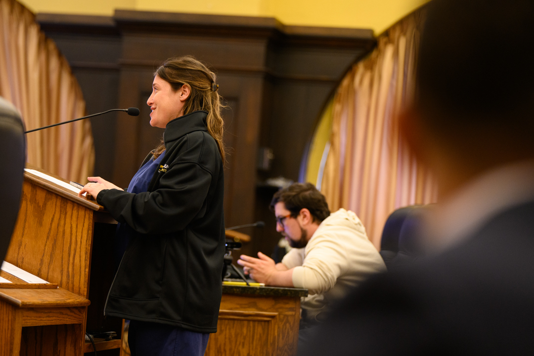 A woman speaks at a podium with a microphone in a room with wood paneling and curtains; a man sits at a desk in the background using a laptop.