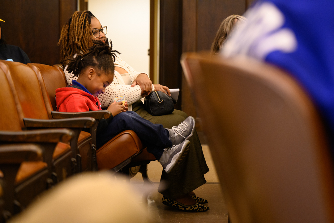 A woman sits in a row of chairs with a child in a red hoodie beside her, who is looking down at an object in their hands.