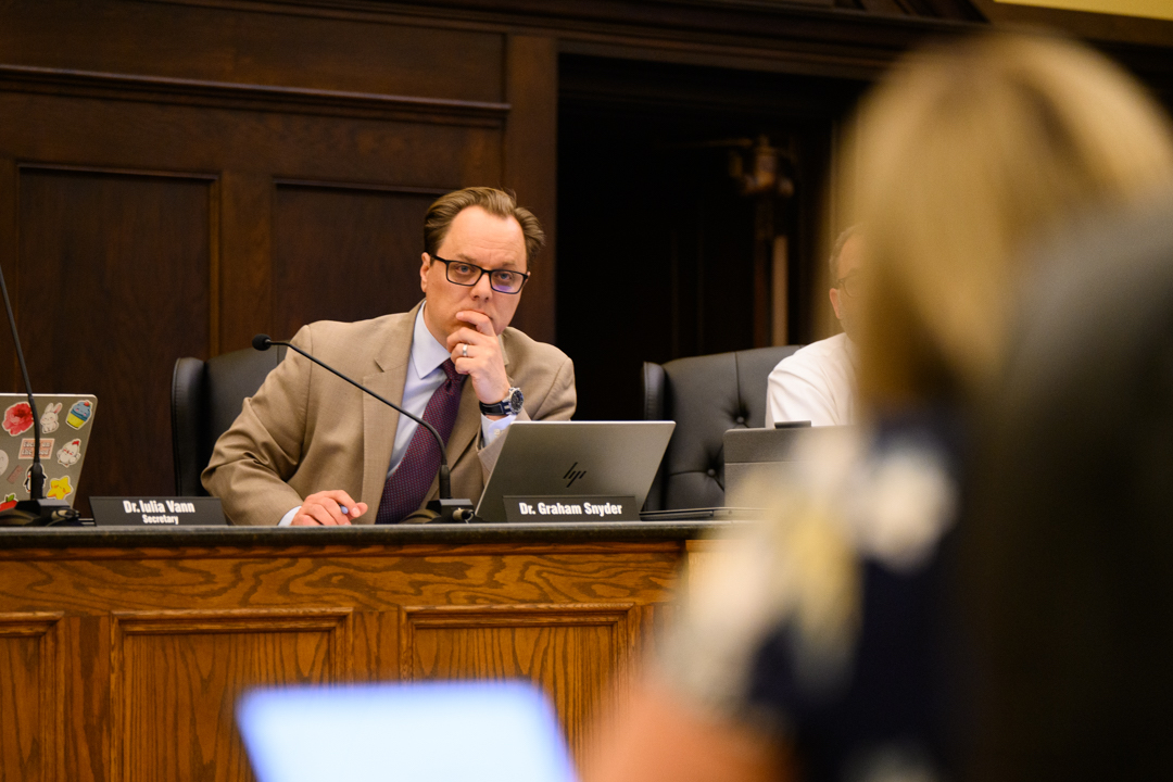 A man in a suit sits at a wood-paneled desk with a laptop, listening attentively during a meeting.