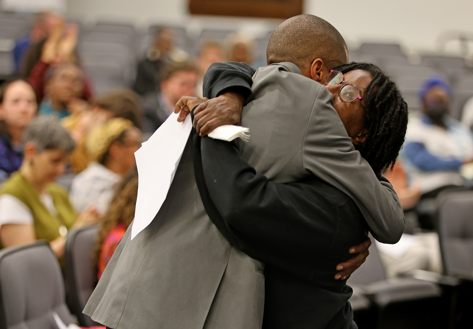 Two people hug in the center of a room with seated attendees in the background, one holding papers, during what appears to be a public meeting or event.