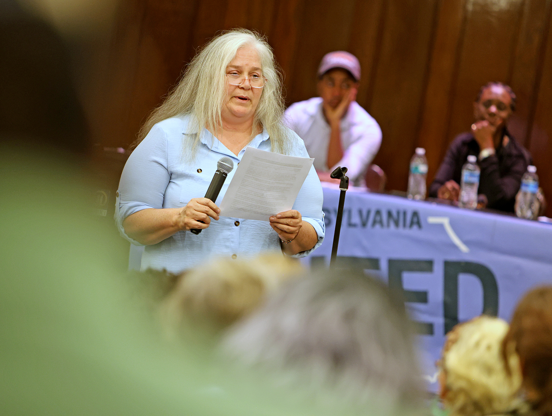 A person with long gray hair reads from a paper while holding a microphone at a public event, with people seated and a blue banner in the background.