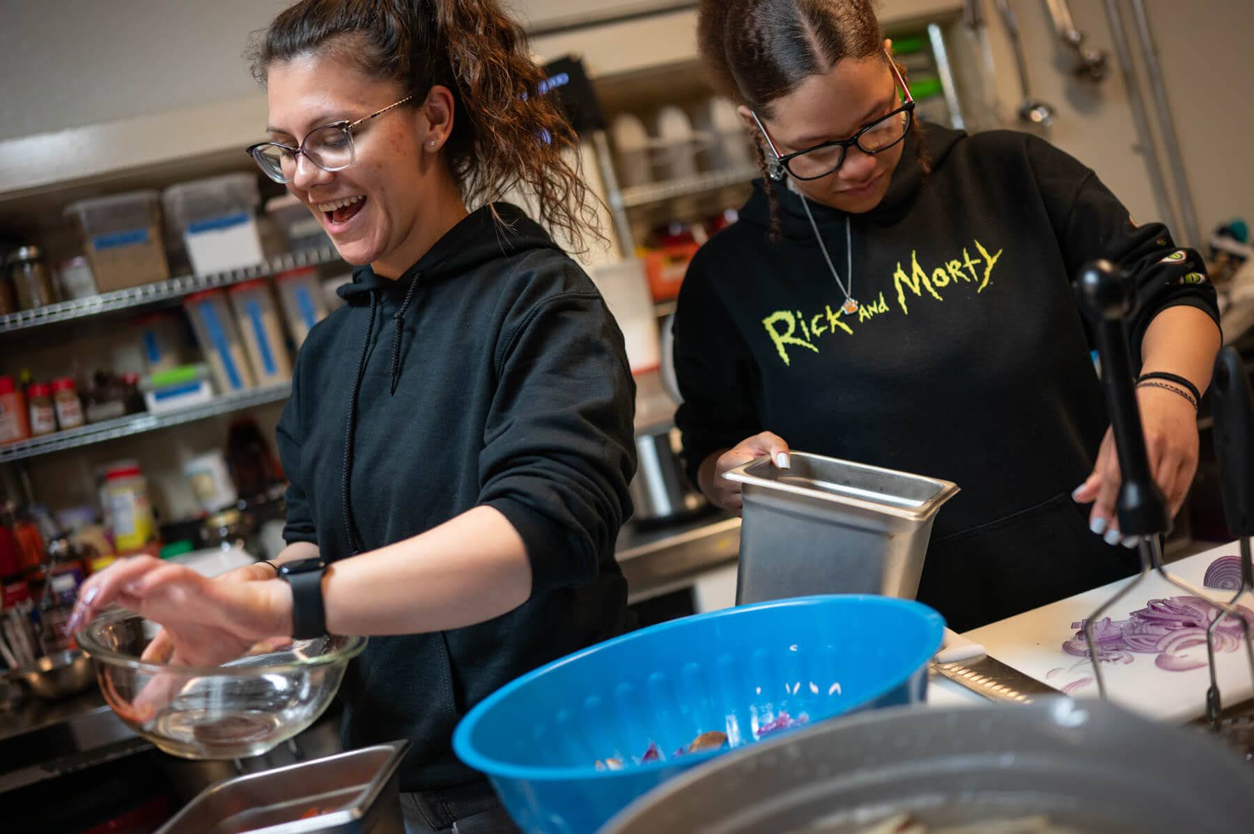 Two people in a kitchen prepare food together. One person is smiling and holding a bowl, while the other is focused on a metal tray. Various kitchen tools and ingredients are visible.