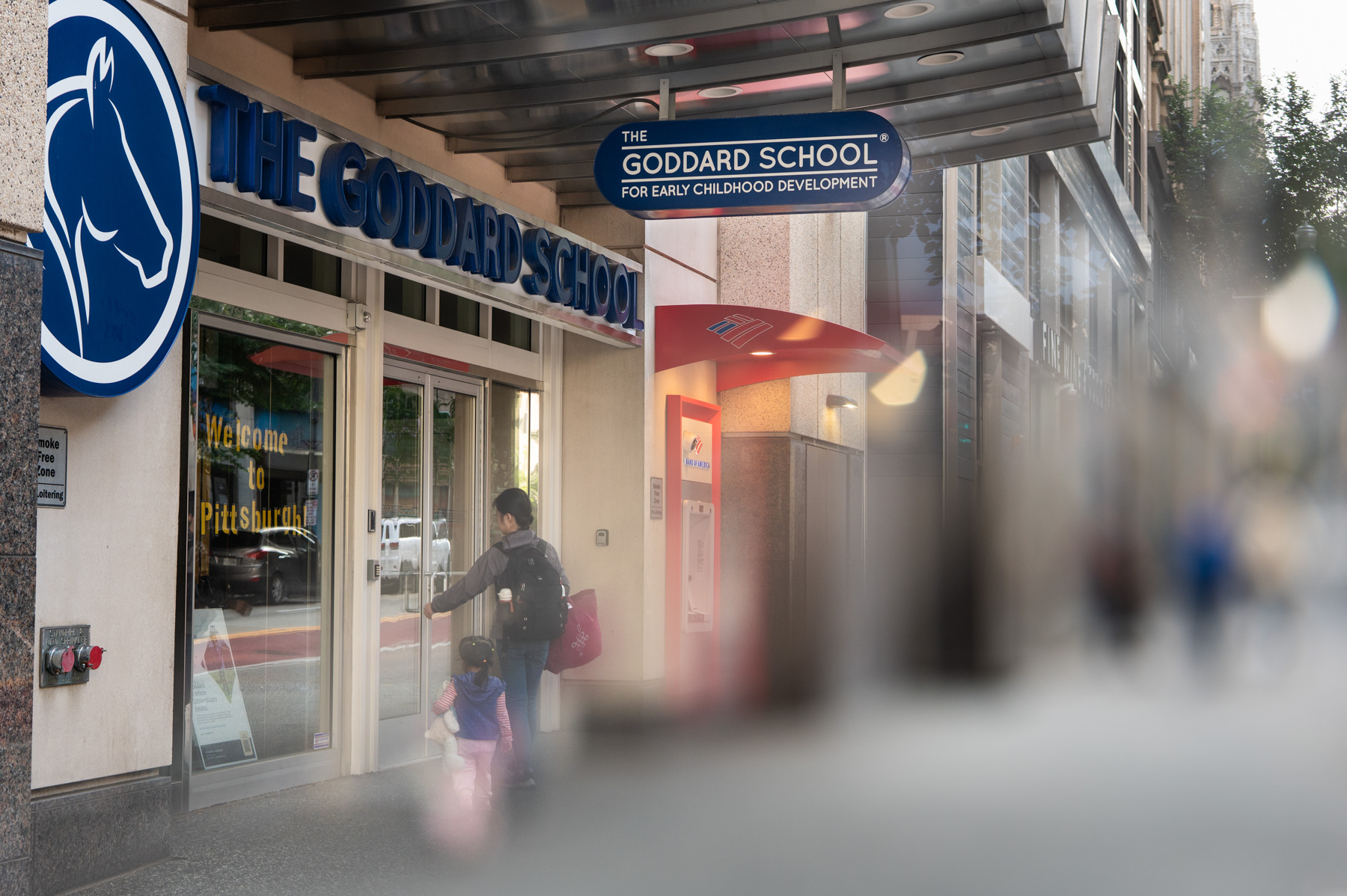 A woman and child enter The Goddard School for Early Childhood Development in an urban area, with a sign reading