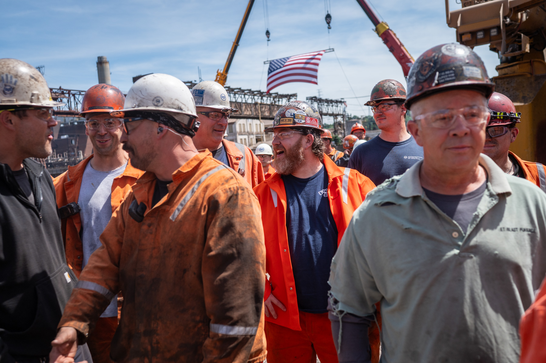 A group of construction workers wearing helmets and safety gear stand together outdoors, with an American flag and industrial equipment in the background.