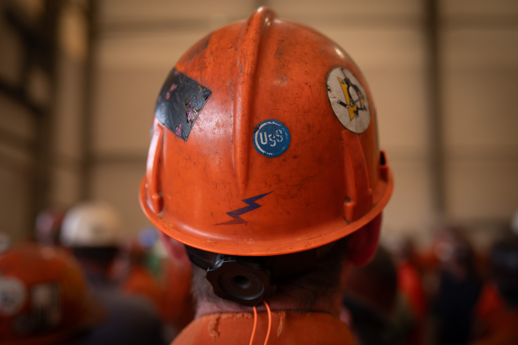 A person wearing an orange hard hat covered in stickers stands among a group of people in an industrial setting.