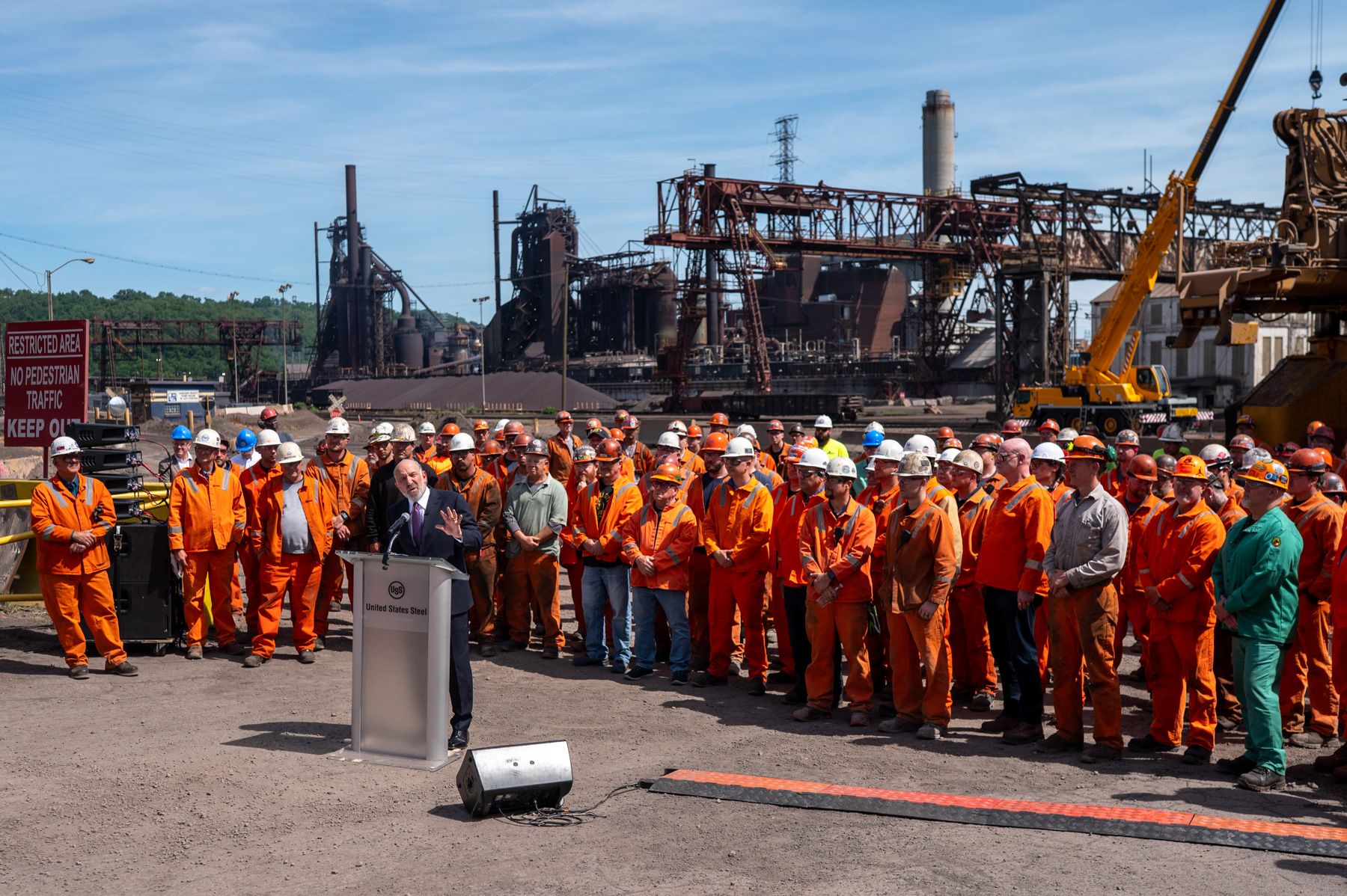 A person speaks at a podium in front of a group of workers in orange uniforms at an industrial site with factories and equipment in the background.