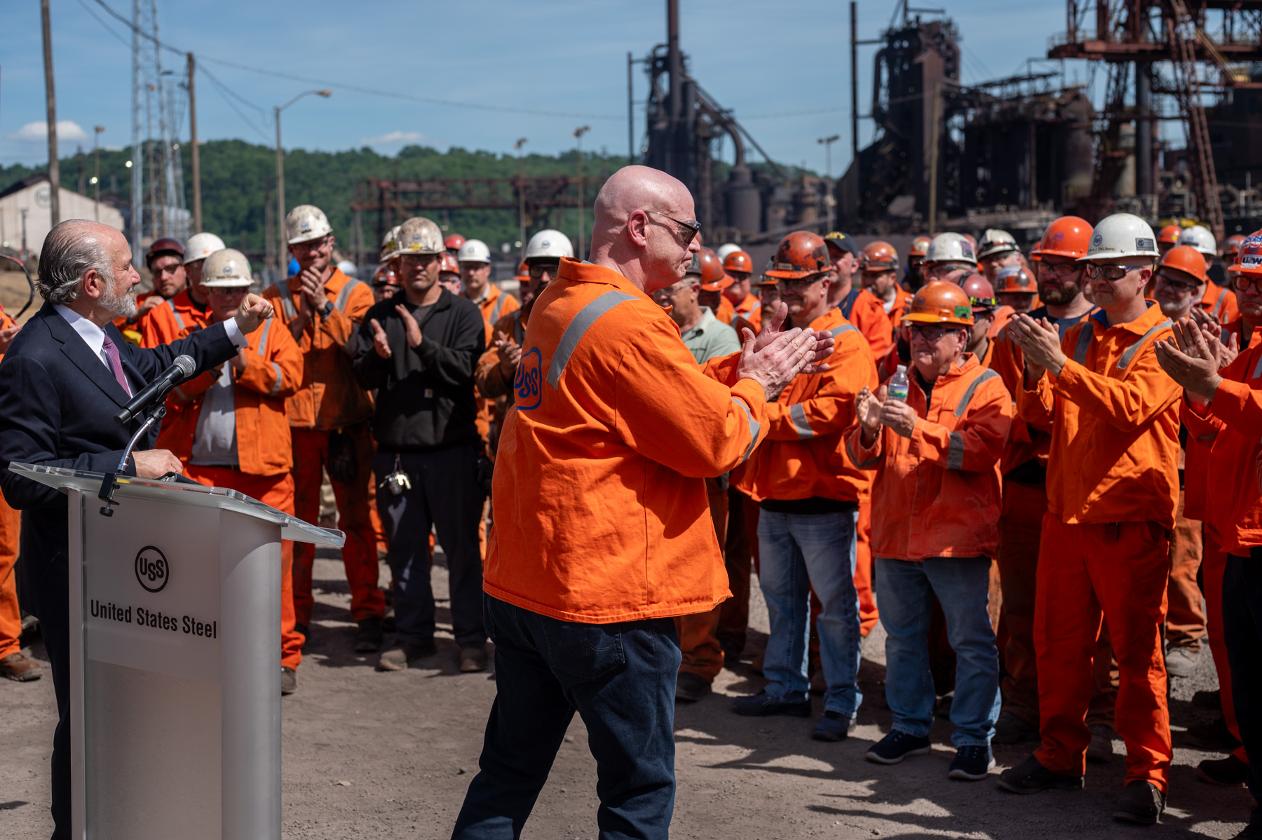 A group of steelworkers in orange uniforms stand and clap near an outdoor podium labeled “United States Steel,” with industrial structures in the background.