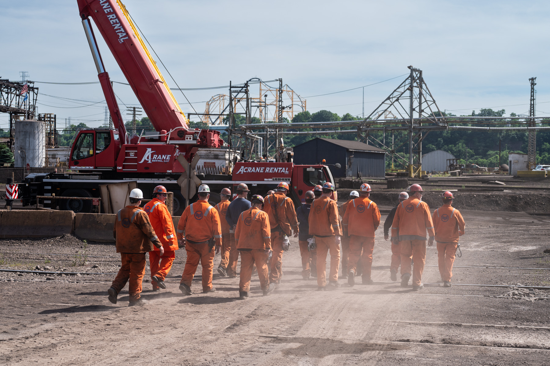 A group of workers in orange safety uniforms and helmets walk towards a red crane labeled 