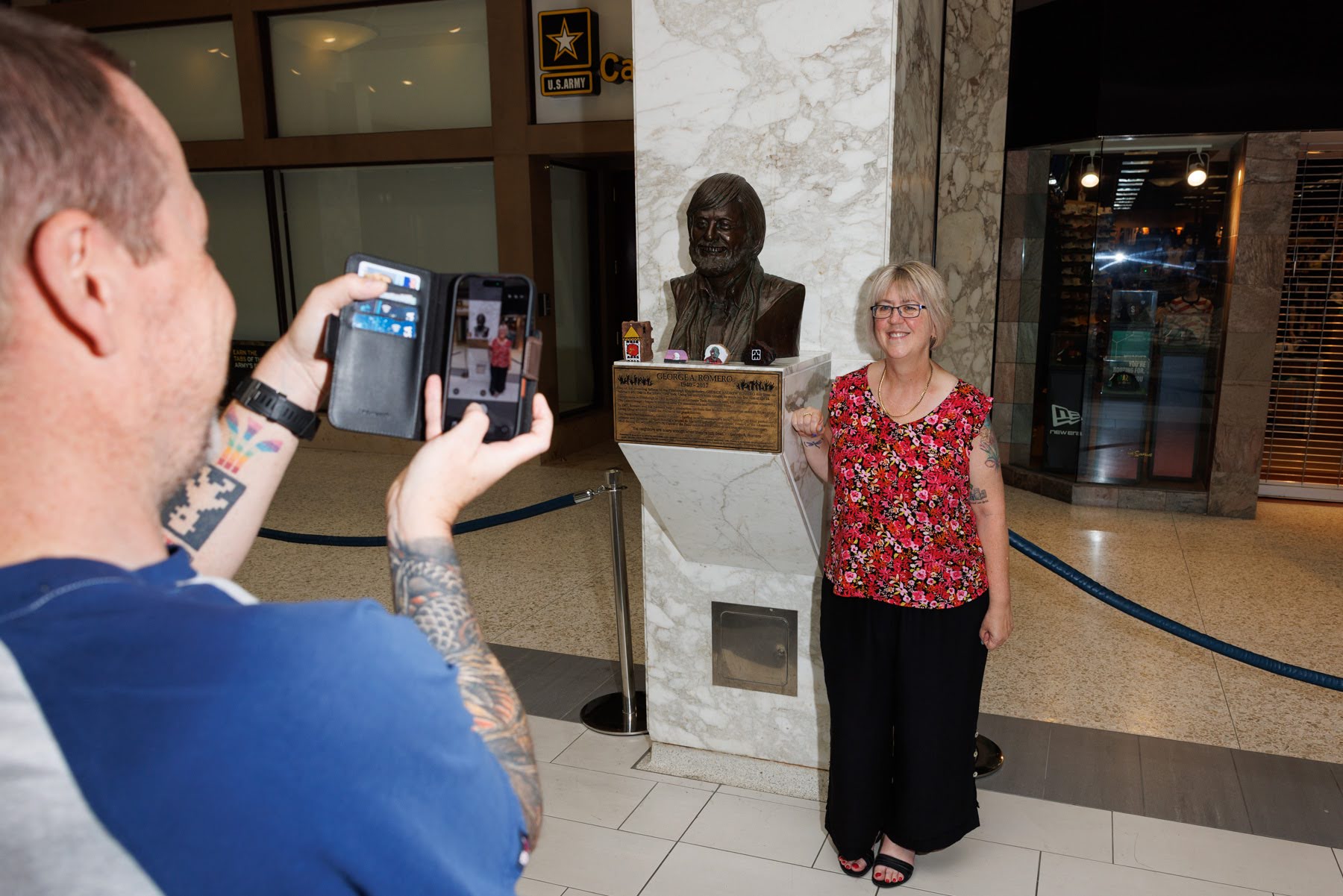 A man takes a photo of a smiling woman standing next to a bronze bust on a marble pedestal inside a mall atrium.