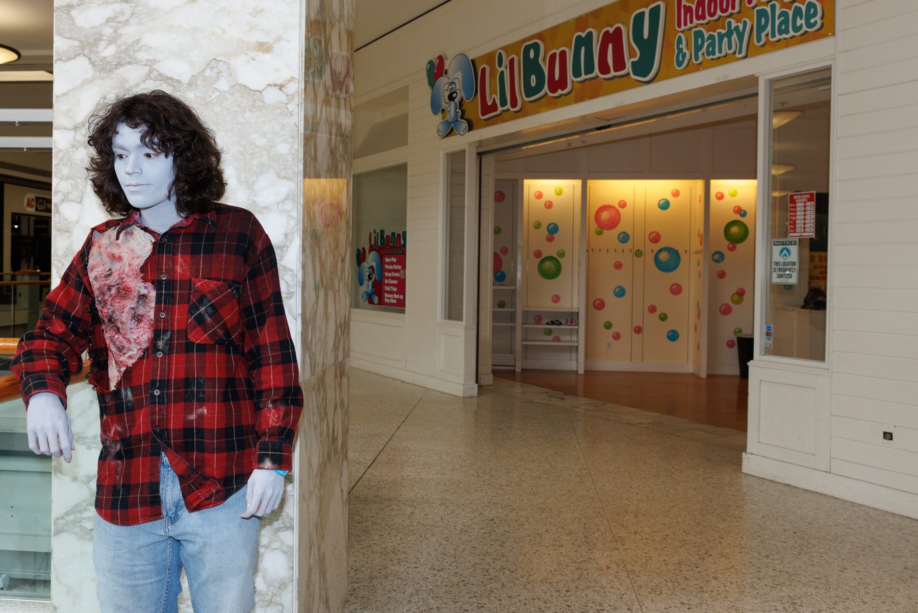 A person dressed in a red plaid shirt and jeans stands by a marble column in a mall hallway near the entrance to