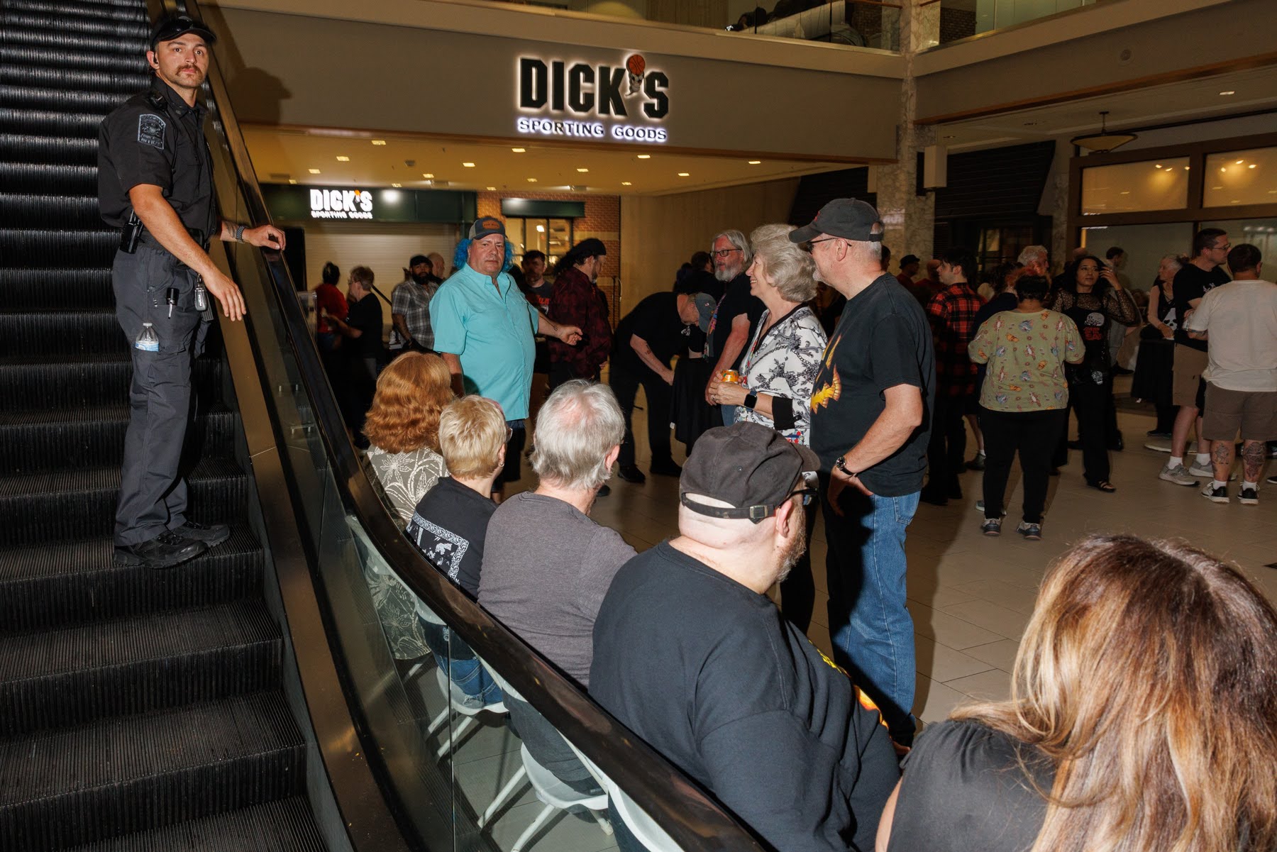 A group of people gathers near the base of an escalator in a shopping mall, with a security guard standing by on the escalator and a Dick's sporting goods store in the background.